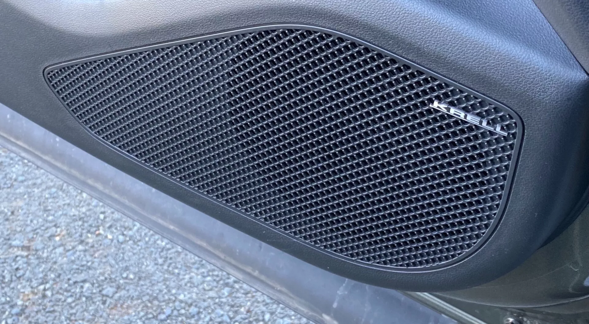 Närbild från sidan av Hyundai Tucson 2020 1.6 T-GDI Pure, med fokus på en sidogaller med ett mönster av diamantskurna nät. Gallret, inneslutet i en formad kontur, är inbyggt i bilens karosspanel med en matt finish, omgiven av en slät, böjd yta. Bilden, tagen i sned vinkel från botten upp, visar naturligt utomhusljus med bitar av asfalt och vägrester, vilket förbättrar det eleganta, prestandaorienterade utseendet.