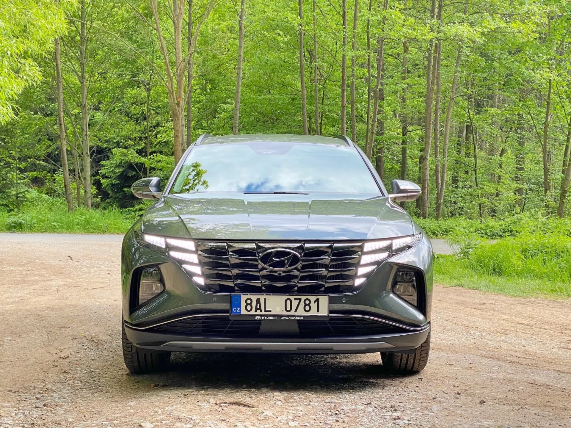 Frontal vy av en Hyundai Tucson 2020 (Hyundai Tucson 1.6 T-GDI Pure) modern SUV parkerad på en grusväg med en skogsbakgrund. Bilden fångar den eleganta, aggressiva designen med en stor geometrisk grill med tillverkarens logotyp, smala vinklade LED-strålkastare och formad främre stötfångare med luftintag och urtag för dimljus, vilket understryker avancerad aerodynamik och belysningsteknik.