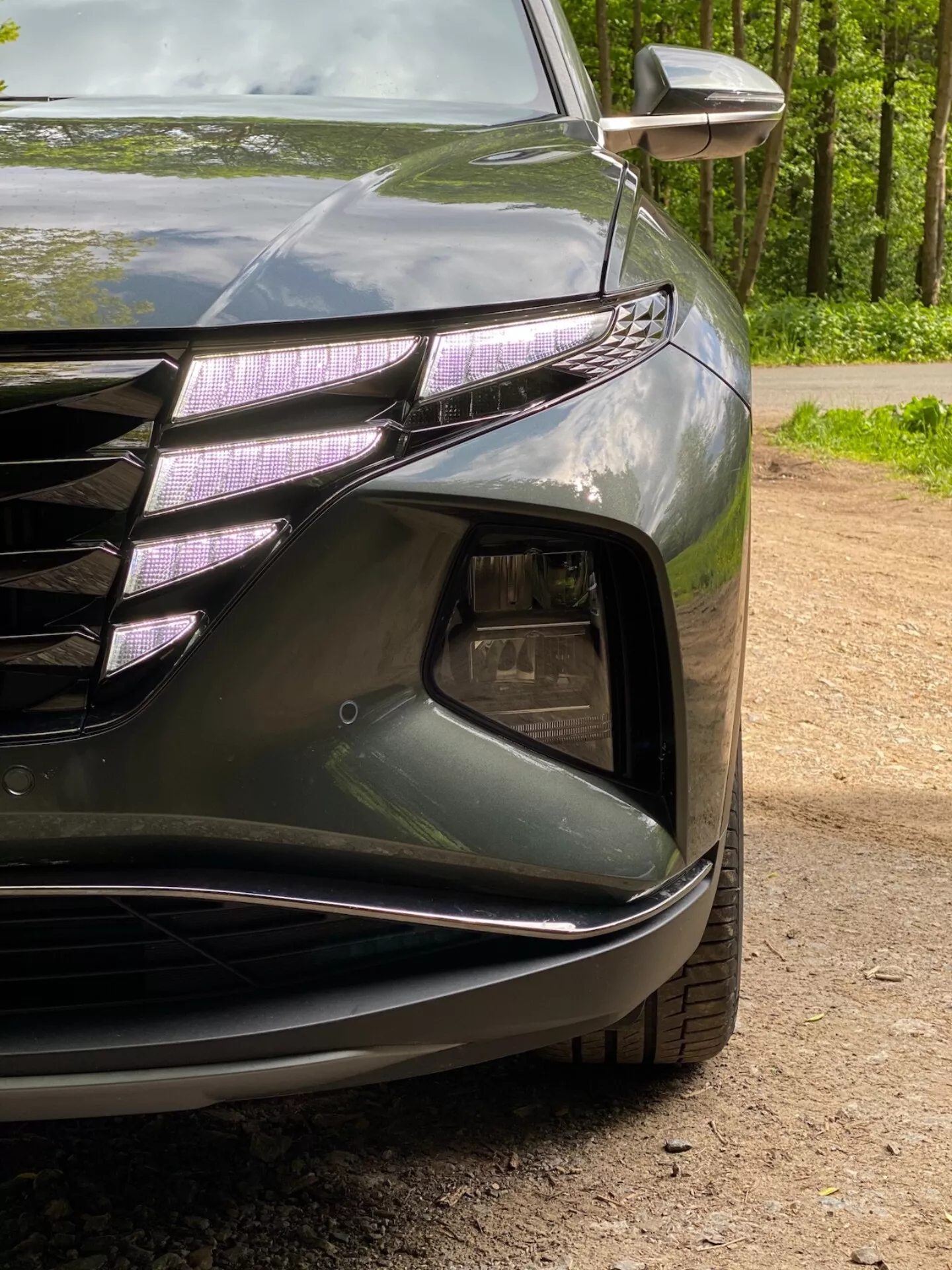 Närbild av det vänstra främre hörnet på en Hyundai Tucson 2020 1.6 T-GDI Pure, som visar strålkastare med LED-dagljus och projektorlins, en del av den främre grillen med horisontella linjer och kromdetaljer, luftintagssektion med dimljus, skulpterad främre stötfångare med släta linjer och integrerad blinkers i ytterbackspegeln. Den mörka, blanka lacken framhäver bilens aerodynamiska kurvor.