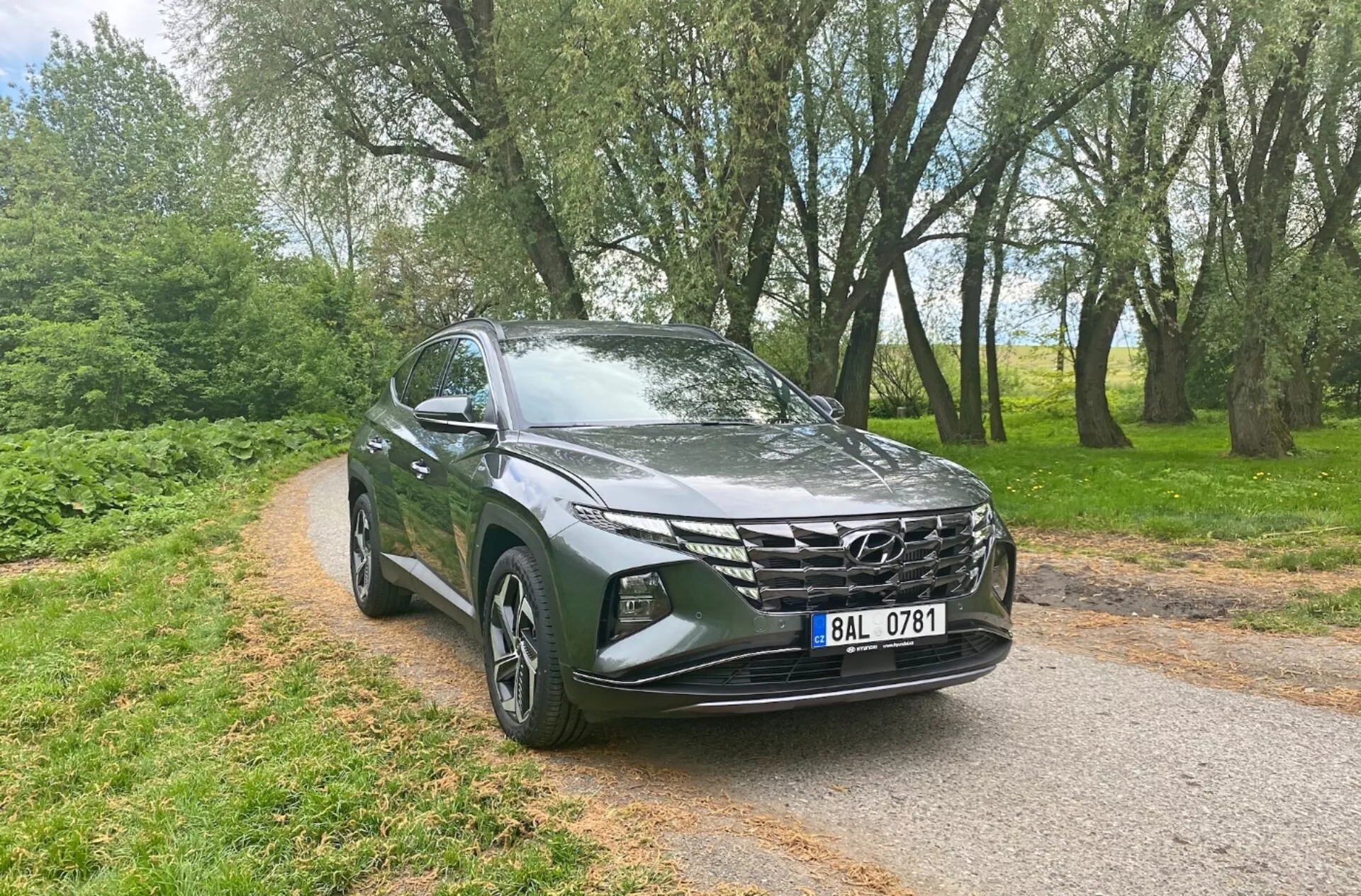 Främre trekvartsvy av en 2020 Hyundai Tucson 1.6 T-GDi Pure, som visar moderna SUV-funktioner som eleganta LED-strålkastare, en framträdande geometrisk frontgrill, skulpterad aerodynamisk huv, lättmetallfälgar, sidobackspeglar med integrerade blinkers och metallisk färg, på en smal asfalterad väg med träd i bakgrunden, vilket betonar dess mångsidighet och attraktionskraft för både urbana och lantliga miljöer.