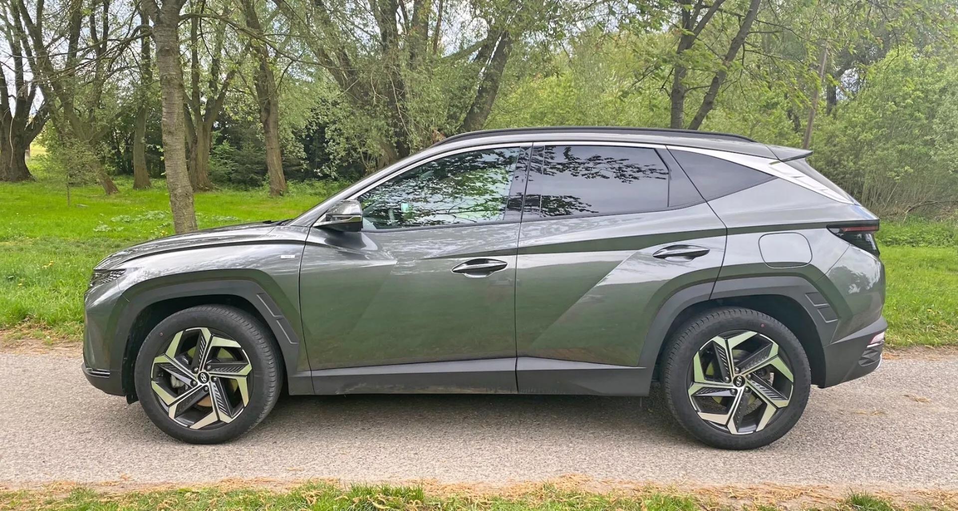 Sidoprofil av en modern 2020 Hyundai Tucson 1.6 T-GDI Pure SUV parkerad på en asfalterad väg bredvid ett grönområde med träd. Visar upp sin eleganta, aerodynamiska design med sluttande taklinje, distinkt karosslinje, stora flerspårs lättmetallfälgar med lågprofildäck, tonade rutor med en synlig B-stolpe, spolade dörrhandtag, karossfärgade sidobackspeglar och kromad trim längs fönsterkanten. Fordonet fångar en samtida stil och möjliga avancerade funktioner ur en sidovinkel.
