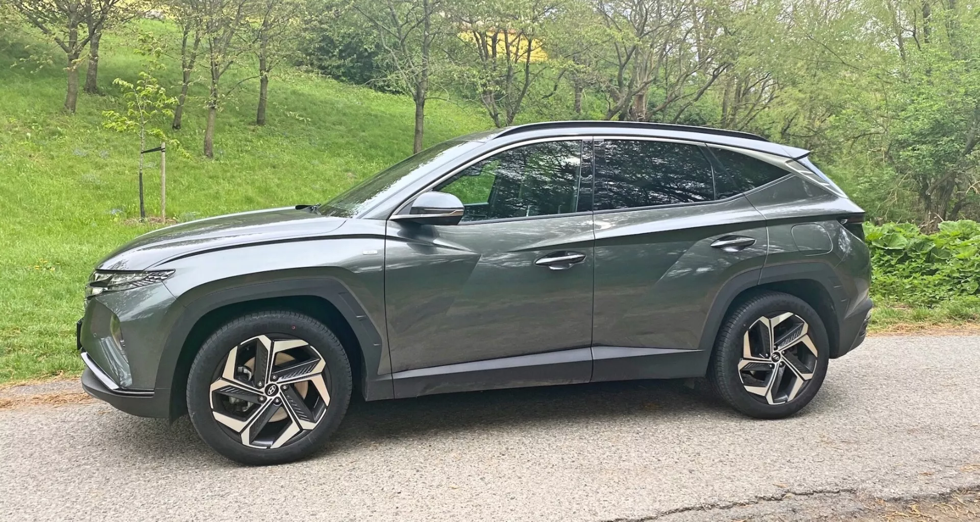 Sidovy av en modern 2020 Hyundai Tucson 1.6 T-GDI Pure crossover SUV i metallicgrå med blank finish. Fordonet har stora, intrikata flerekerfälgar, LED-strålkastare, lätt tonade fönster, aerodynamiska karosslinjer med en sluttande taklinje som slutar i en bakre spoiler, takräcken, sidospeglar med integrerade blinkers och krom- eller metallaccenter. Fordonet är placerat på en asfalterad väg med en lätt nedåtgående vinkel och placerat mot höger i bilden.