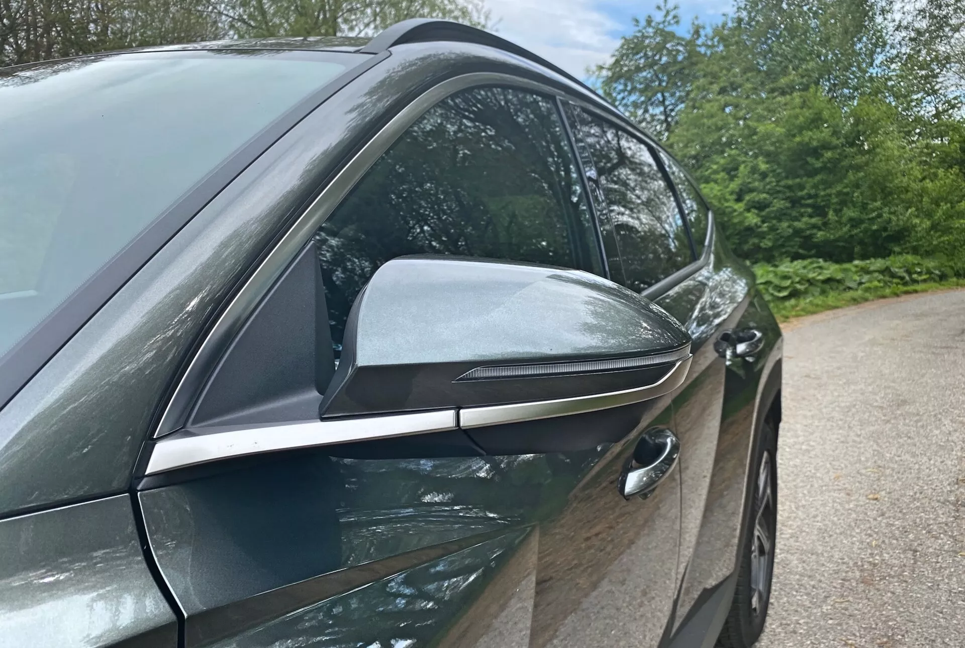 Närbild av sidan på 2020 Hyundai Tucson 1.6 T-GDI Pure som framhäver dess moderna design och tekniska funktioner med fokus på förarsidan. Bilden tagen från låg vinkel visar den eleganta, aerodynamiska sidobackspegeln med integrerade blinkers och svarta accenter, vilket antyder avancerade funktioner. Den infällda förardörrshandtaget antyder ett nyckellöst instegssystem, vilket bidrar till bilens aerodynamiska effektivitet. De glänsande, bilfärgade dörrarna och kromfönsterlister antyder ett premium trim-paket. Bakgrundselement som träd och himmel antyder en utomhusmiljö.