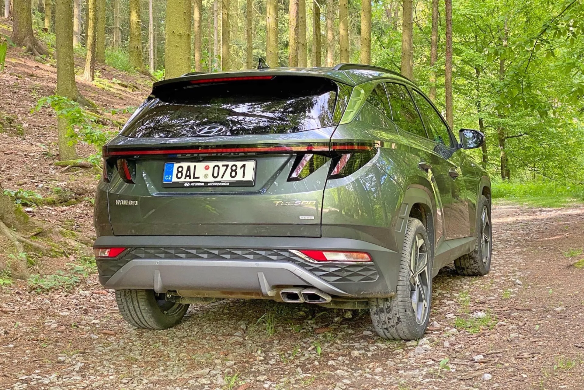 Bild av en modern Hyundai Tucson 2020 SUV i en grusig skogsmiljö, tagen från en bakre trekvartsvinkel. Fotot lyfter fram fordonets högra sida och bakprofil med synlig baklucka, LED-bakljus, dubbla avgasrör, bakspoiler och lättmetallfälgar.