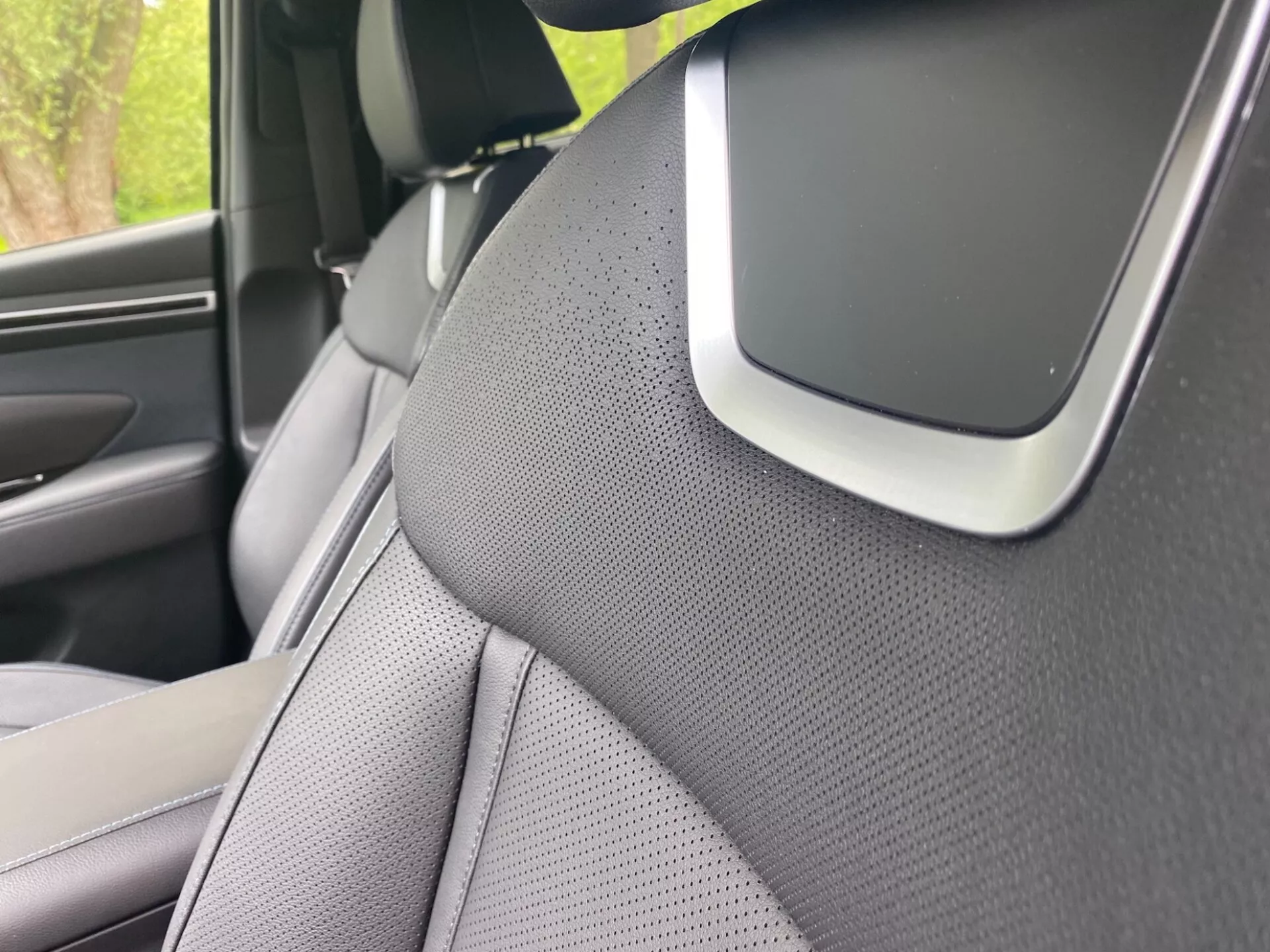 Närbild av interiören i Hyundai Tucson 2020 med fokus på den högra A-stolpen och en del av den högra framsätet. A-stolpen är klädd med ett texturerat material, troligen tyg eller konstläder, vilket antyder fokus på estetik eller bullerdämpning. Sätet har en tvåfärgad design och är perforerat för ventilation. En märkbar funktion vid korsningen av A-stolpen och innertaket kan vara en del av ljudsystemet, en airbag-komponent eller en förarassistanssensor, vilket framhäver avancerade säkerhets- eller teknologifunktioner.