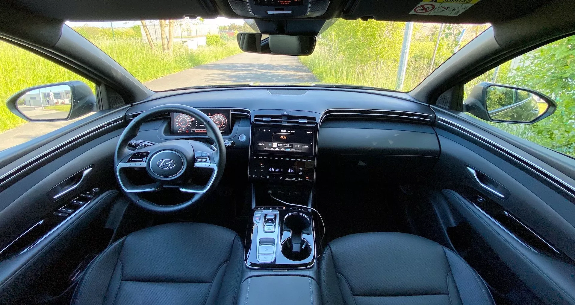 Interiörvy av Hyundai Tucson 2020 från baksätet som visar framinstrumentpanelen, ratt med monterade kontroller, digital instrumentpanel, stor pekskärmsinfoentertainmentsystem, klimatkontrollknappar, växelspak och framsäten klädda i läder under naturlig dagsbelysning.