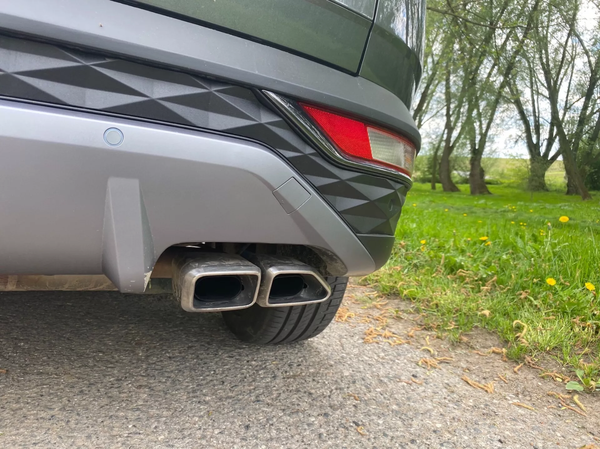Närbild av bakre hörnet på en Hyundai Tucson 2020, tagen från marknivå. Synliga detaljer inkluderar en tvåfärgad bakre stötfångare, en delvis LED-baklampa och dubbla metalliska avgasrör med rektangulära spetsar, vilket tyder på ett sportigt trim. Fordonets eleganta design och metalliska lack reflekterar den naturliga omgivningen.