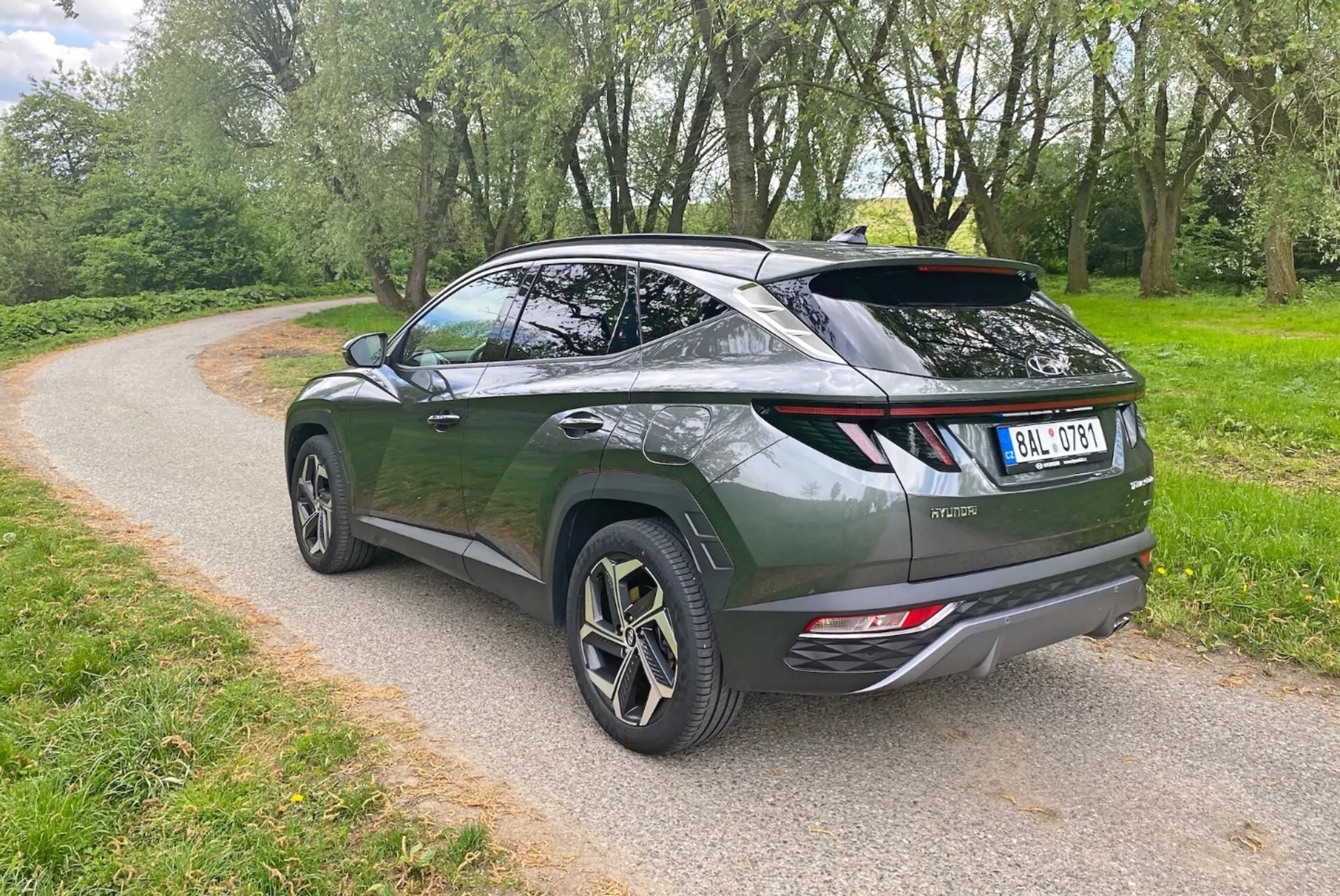 Bilden visar en modern crossover SUV från en trekvarts bakvinkel, som framhäver den eleganta, aerodynamiska designen och den sluttande taklinjen. Det mörkgröna fordonet har en baklucka i liftback-stil, sportig spoiler och stora legeringshjul med en flerspoked design. Synliga element inkluderar moderna bakre LED-lampor, dubbla avgasrör och en bakre vindrutetorkare. Suven är parkerad på en grusstig omgiven av grönska, vilket tyder på en park- eller lantlig miljö.