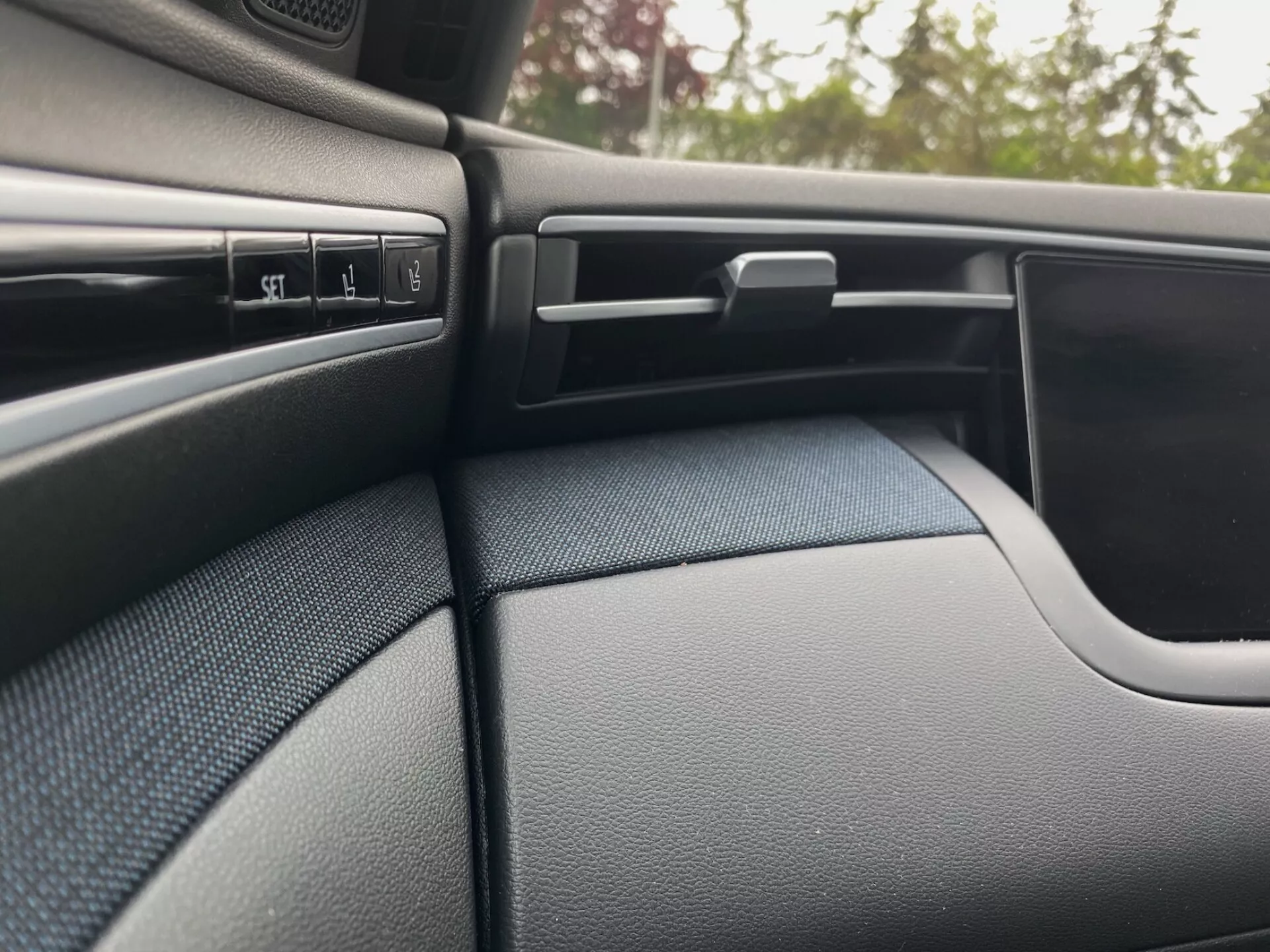 Interiördetalj av Hyundai Tucson 2020, som visar den mörkfärgade, texturerade passagerarsidan av instrumentpanelen och delen av dörrpanelen. Scenen framhäver flera kontrollknappar, en justerbar ventilationsöppning med horisontella lameller, ett låsbart handskfack och den kurviga designen av dörrpanelen som smälter sömlöst in i instrumentpanelen.