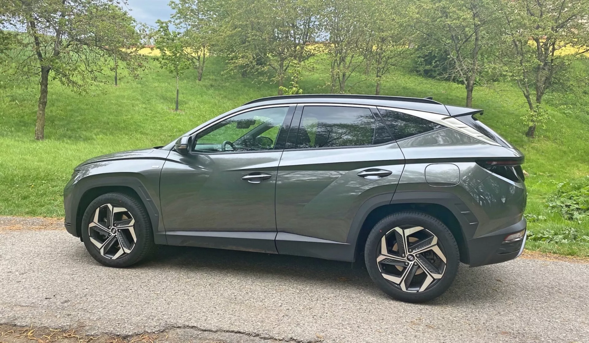 Sidovy av en Hyundai Tucson 1.6 T-GDI Pure 2020 SUV i naturligt dagsljus. Fordonet är placerat i ett grönområde med träd i bakgrunden, vilket antyder en lantlig eller förortsmiljö. Moderna designegenskaper inklusive LED-strålkastare, integrerade sidospegelsignaler, tonade rutor, aerodynamiska lättmetallfälgar och en kupéinspirerad sluttande taklinje syns. Den släta karossen med karaktärslinjer och dolda dörrhandtag antyder ett avancerat nyckellöst instegssystem, medan den bakre spoilern indikerar aerodynamiska förbättringar. Bilen är centralt placerad i ramen och ger en tydlig, obehindrad vy från medelhöjdsperspektiv.