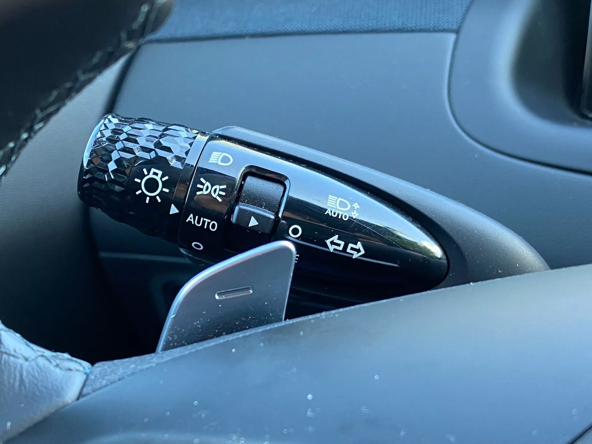 Närbild av kontrollspaken belägen på rattstången till vänster om ratten i en Hyundai Tucson 2020. Spaken har symboler för strålkastarinställningar inklusive av, automatiska strålkastare, parkeringsljus och halv-/helljusstrålkastare. Bilden fångar spaken, en del av ratten och en del av förardörren i suddig bakgrund, med fokus på detalj och designkvalitet. 'AUTO'-positionen indikerar ett automatiskt strålkastarsystem.