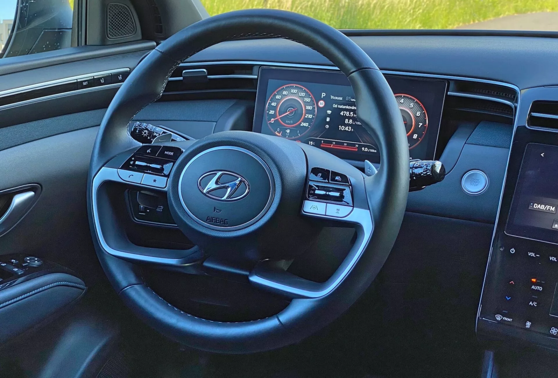 Interiörvy av Hyundai Tucson 1.6 T-GDI 2020 från förarens perspektiv, med fokus på den moderna treekrade ratten med Hyundai-logotyp, media- och farthållarknappar, delvis synlig instrumentbräda med hastighetsmätare och varvräknare, samt central infotainmentsystem med pekskärm på instrumentbrädan.