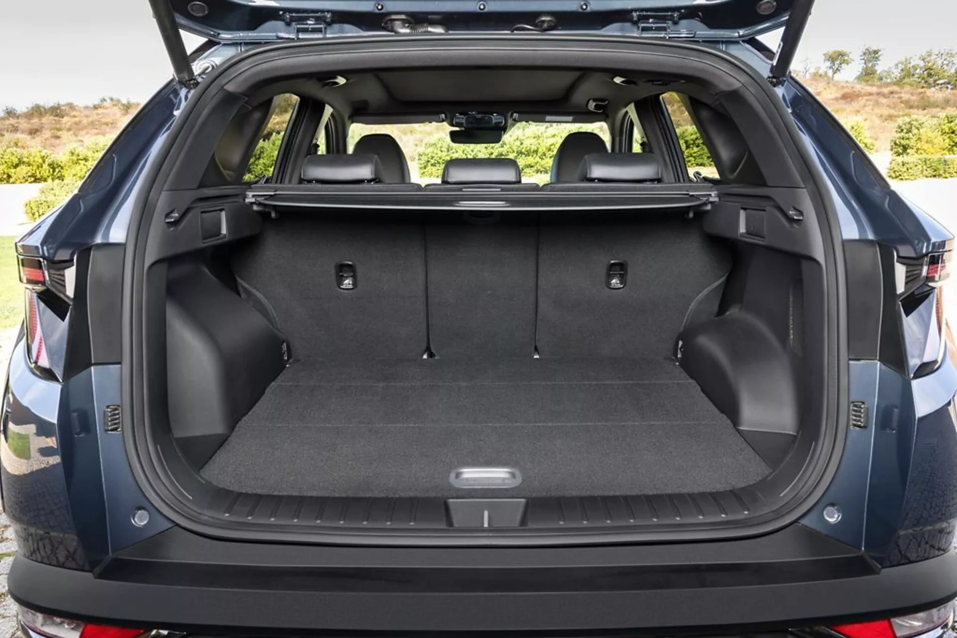 Bakre lastutrymmet i Hyundai Tucson 2020 med bagageluckan öppen, visar upp mattbelagd lastgolvyta, fästpunkter, förvaringsfack, 12V-uttag, fällbara baksäten, indragen lastskydd, tåliga plastsidoväggar och en glimt av bakre stötfångaren, utomhus.