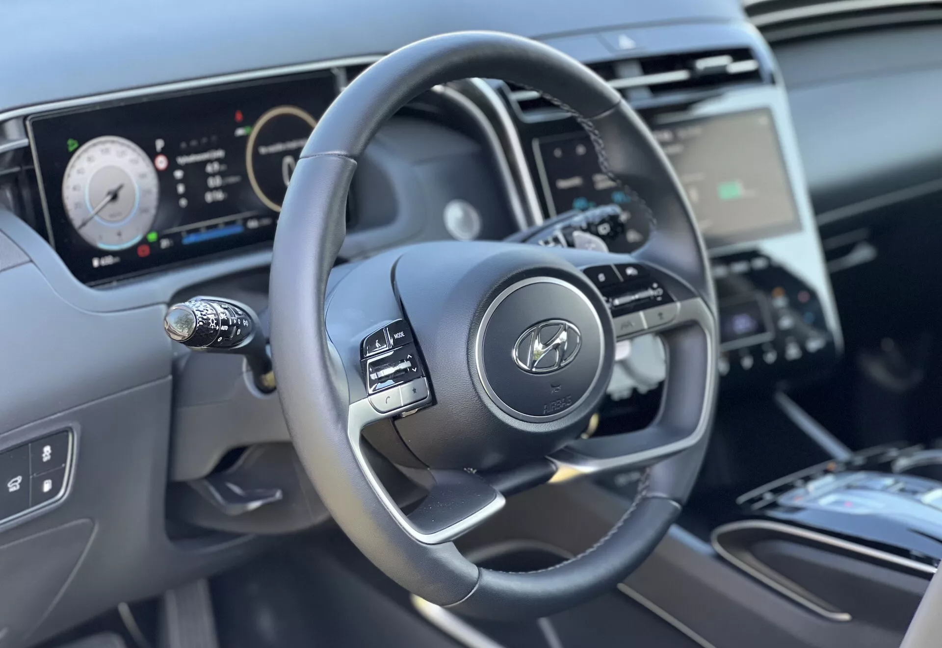 Interiörvy av Hyundai Tucson 2020 med fokus på ratten och instrumentbrädans funktioner från förarens perspektiv, framhäver den trespekiga designen med kontrollknappar, digital instrumentkluster och delvis vy av mittenkonsolen och klimatkontroll.