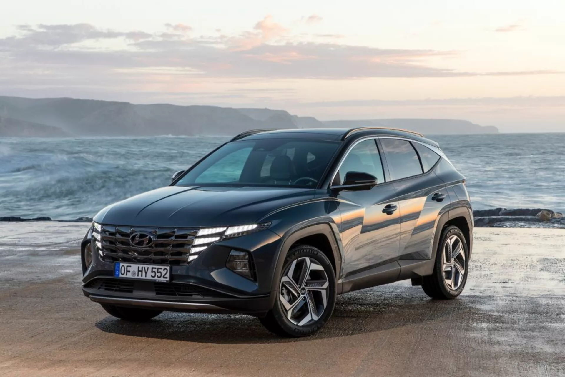 Modern SUV Hyundai Tucson 2020 parkerad på en strand med havet och horisonten i bakgrunden, visar dess främre tre kvartalsvy som framhäver fordonets design, framträdande grill med logo, vinklade LED-strålkastare och sportiga legeringshjul