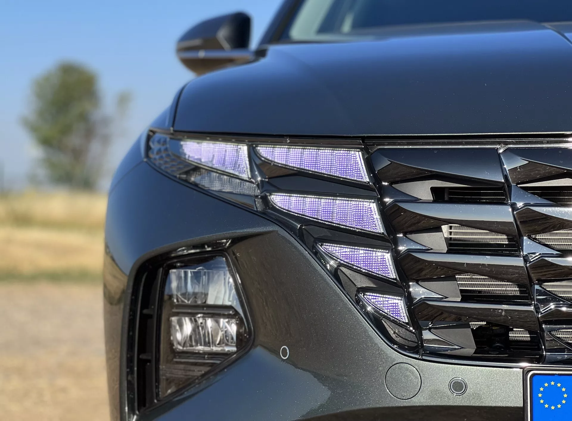 Närbild av den främre vänstra kvadranten på Hyundai Tucson 2020 som visar LED-strålkastare och en detaljerad grill med kromfinish mot en oskarp utomhusbakgrund
