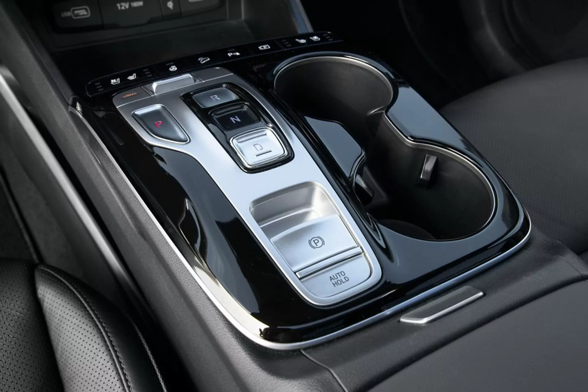 Närbild av mittkonsolen i en Hyundai Tucson 2020 som visar växelspak för automatväxellåda, kontrollknappar, Auto Start/Stopp-funktion, varningsljusomkopplare och dubbla dryckeshållare med kromdetaljer, ur ett översiktsperspektiv med lätt lutning.