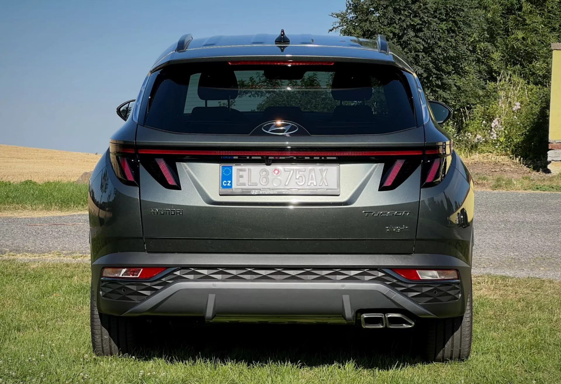 Baksida av Hyundai Tucson 2020 som visar horisontella bakljus, central emblem, trimningsnivånamn, integrerade parkeringssensorer på stötfångaren och ett dubbla avgassystem, parkerad på en gräsbelagd yta mot en klar himmel