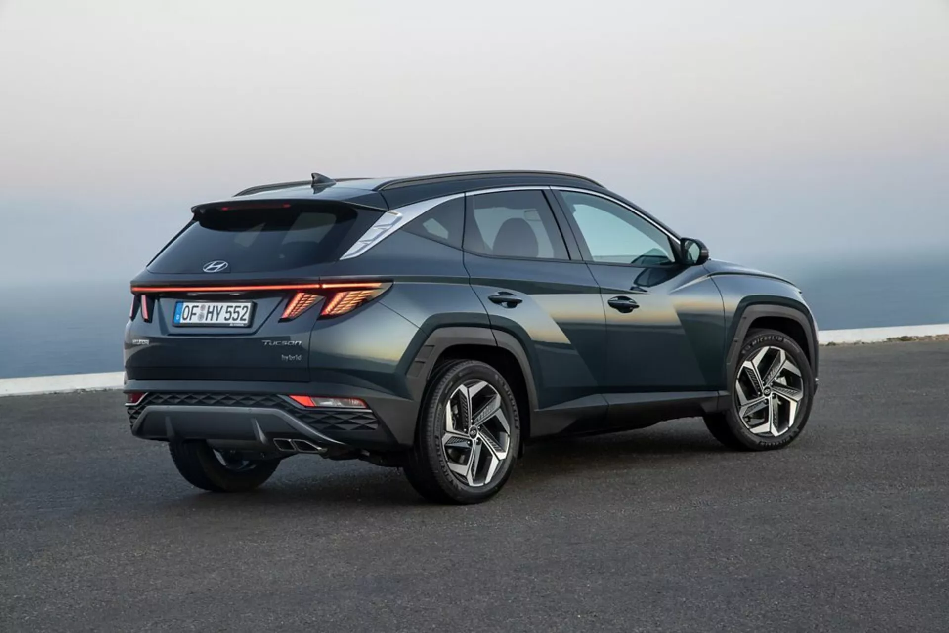 SUV Hyundai Tucson 2020 parkerad på slät asfalt med en kust- eller höjdlandskap i bakgrunden, som visar sin elegant, sportig design, stigande midjelinje, sluttande taklinje, skarpa karosserisveck, vinklade LED-baklampor, bakre spoiler, integrerad diffusor, dubbla avgasrör och stora legeringshjul