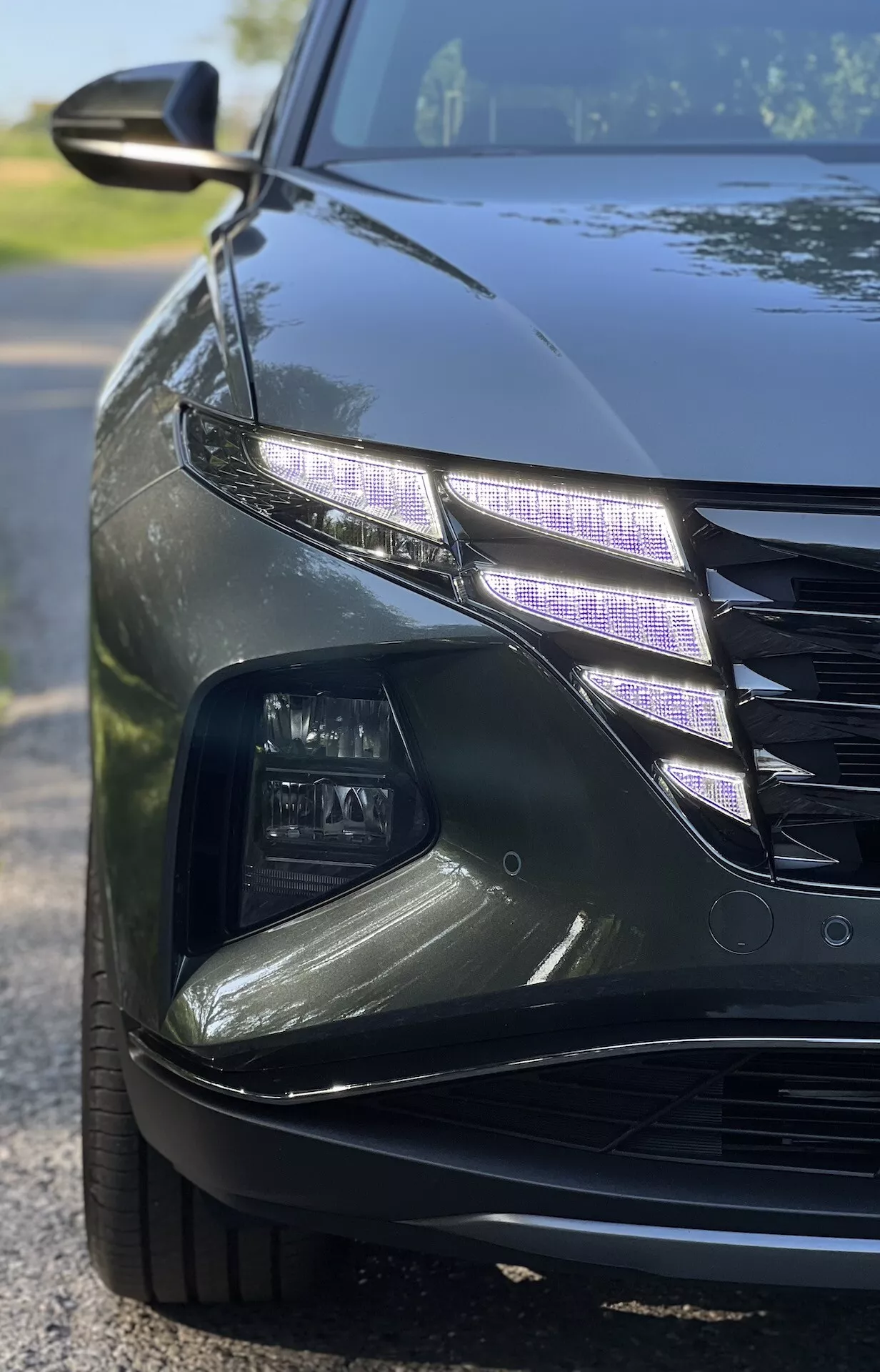 Närbild i vinkel av en Hyundai Tucson 2020 som visar avancerad LED-strålkastardesign och en del av grillen med en glansig finish under svagt ljusförhållanden, vilket illustrerar funktioner i lyxbilar och modern fordonsbelysningsteknik.