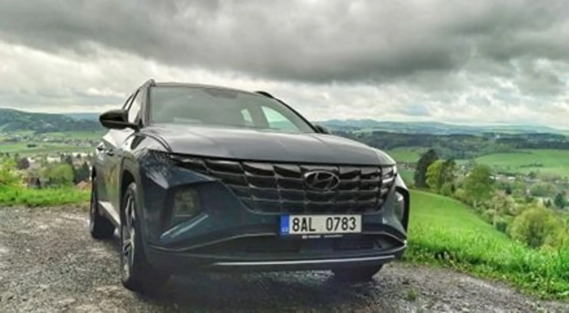 SUV Hyundai Tucson 2020 parkerad på en grusväg, visar upp sin djärva grill, eleganta LED-strålkastare och metallisk lack i en dynamisk vy från en låg främre vinkel, med ett frodigt kuperat landskap under en molnig himmel i bakgrunden