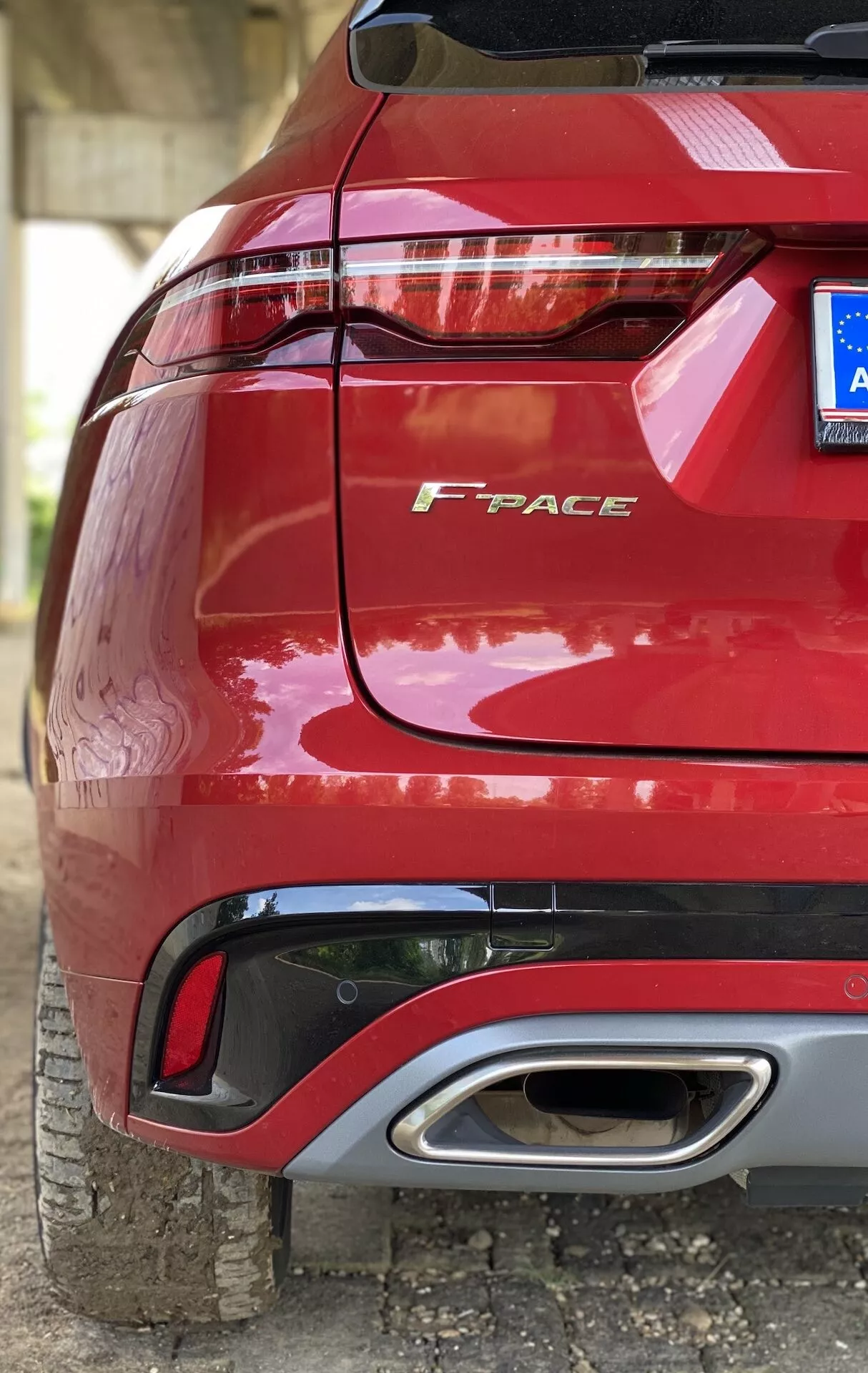 Närbild, vinklad vy av bakdelen på en röd Jaguar F-PACE 2021 som visar vänster sida med fokus på den nedre halvan, fångar bakre stötfångaren, stilfull LED-baklykta, modellnamnet 'F-PACE' i krombokstäver, plastbeläggning, diffusorelementet, ett enda ovalt avgasrör och delvis synligt bakdäck, parkerat på jordig yta.