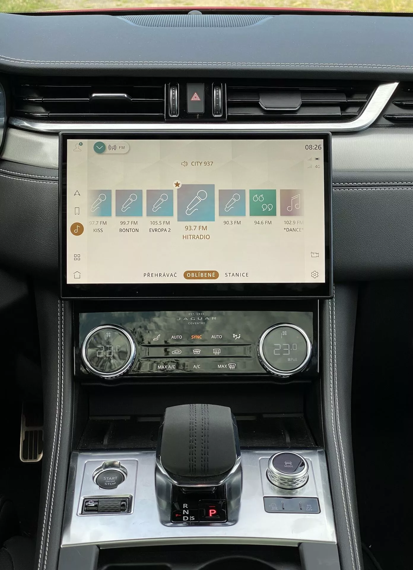 Interiörbild av Jaguar F-Pace 2021 som fokuserar på mittkonsolen och instrumentbrädan, med en horisontellt orienterad pekskärm för infotainmentsystem med navigering, media, telefonanslutning och inställningar. Skärmen visar tiden 08:26, signalstyrka, 17°C temperatur och under finns en varningsknapp och luftventiler. Två analoga rattar, möjligen för klocka och en timer eller kompass, kontrasterar mot den moderna skärmen. Den elektroniska läderväxelspaken är i 'Park'-läge, omringad av körlägesväljare och ett litet förvaringsfack med ett 12-voltsuttag. Den lyxiga interiören kombinerar svarta material med silverdetaljer.
