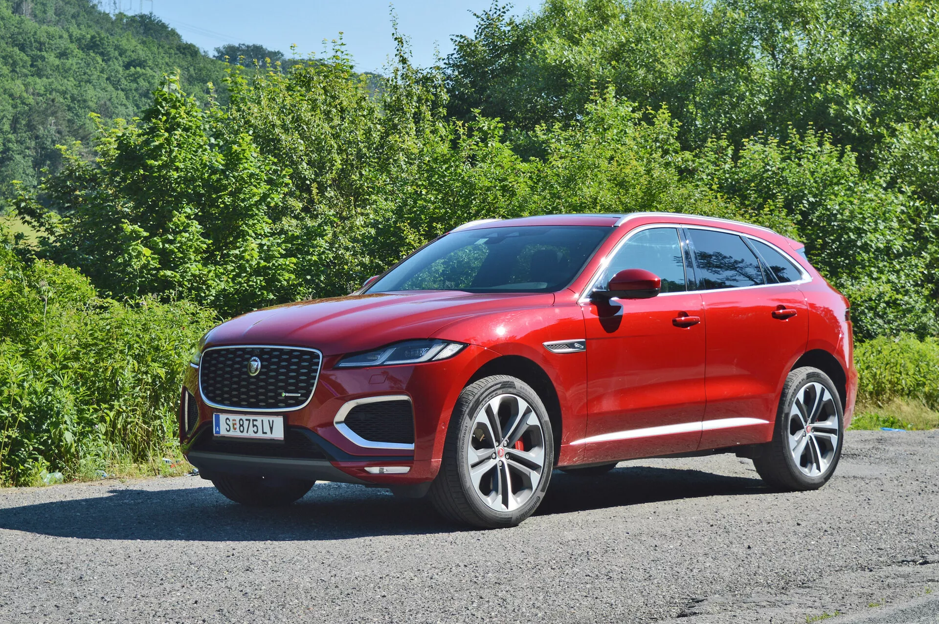2021 års röd Jaguar F-Pace parkerad på en smal väg med vegetation i bakgrunden. Bilden är tagen från en tredjedels frontvinkel som visar både front- och sidoprofilerna. Kännetecknas av den framträdande frontgrillen med ett nätmönster, eleganta LED-strålkastare, synliga parkeringssensorer på frontstödet, stora fälgar i flera ekrar, aerodynamiska karosslinjer, kroppsfärgade sidospeglar med integrerade blinkers och takräcken för extra lastkapacitet.