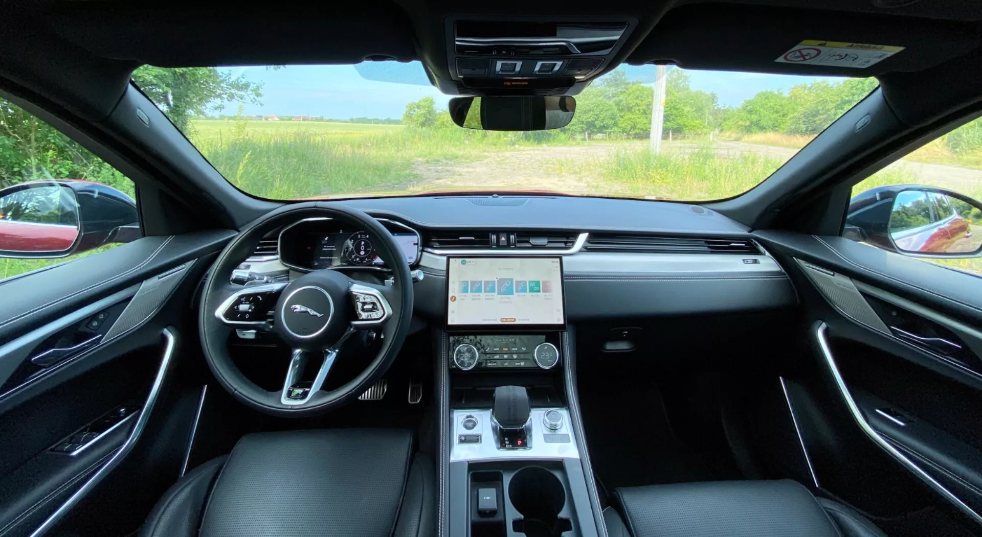 Interiörvy från förarsätet i 2021 års Jaguar F-Pace, med en modern, multifunktionell ratt med medie- och farthållarknappar, central digital instrumentpanel, stor pekskärm för infotainmentsystem, ventilationshål, klimatanläggning, automatisk växelspak samt lyxiga lädersäten med metalliska och kromade accenter. Vindrutevyn visar en klar dag med ett grönt område av träd och gräs.