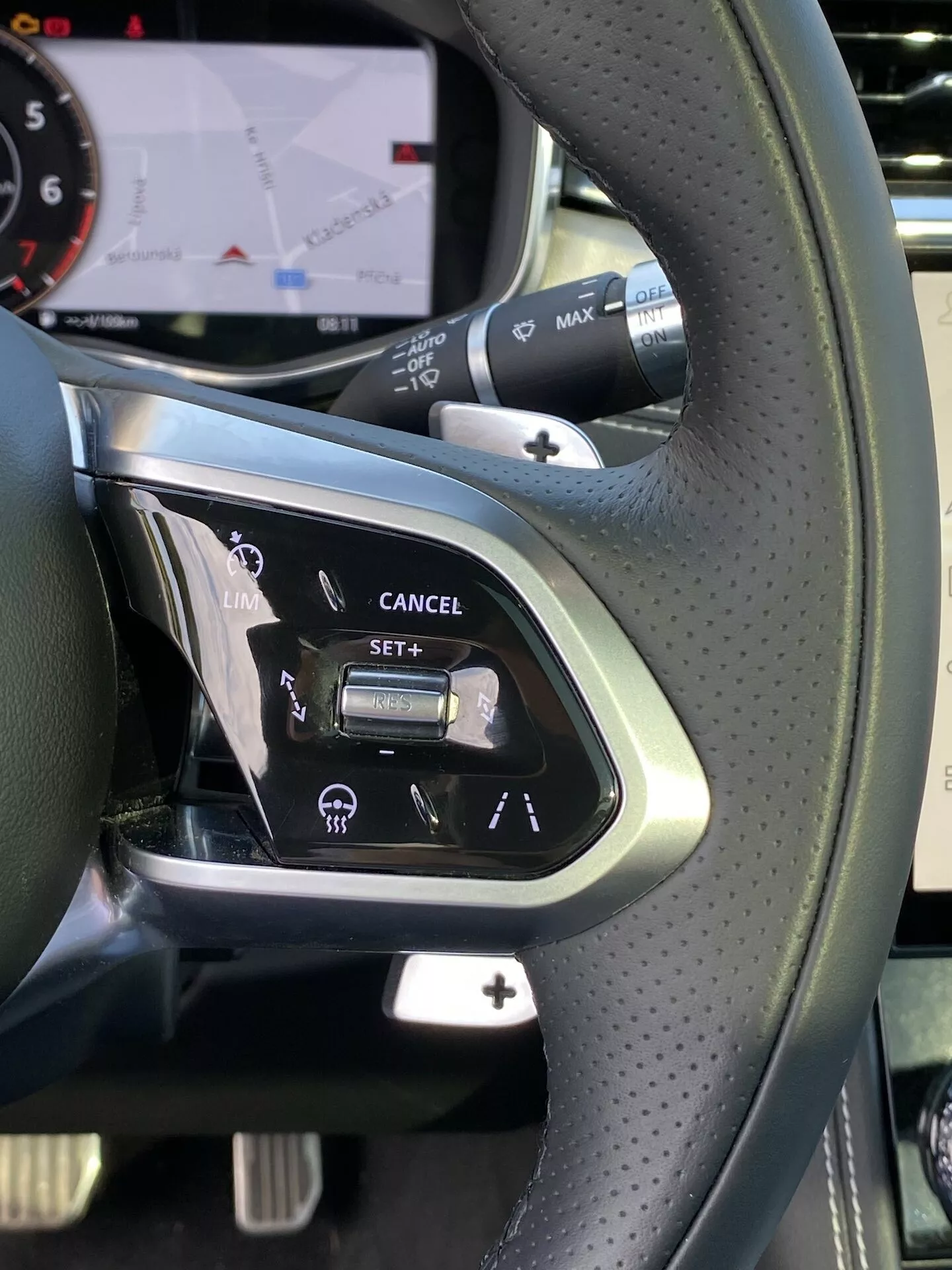 Närbild av vänstra sidan av ratten inuti en lyxbil av modell Jaguar F-Pace 2021, med läderklätt perforerat grepp, silveraccent, farthållarknappar märkta 'LIM', 'CANCEL', 'SET' och en vippströmbrytare, avancerade körhjälpmedel som hastighetsbegränsare och filhållningsassistans, röstkommandoknapp och del av en digital instrumentpanel med navigeringskartskärm.
