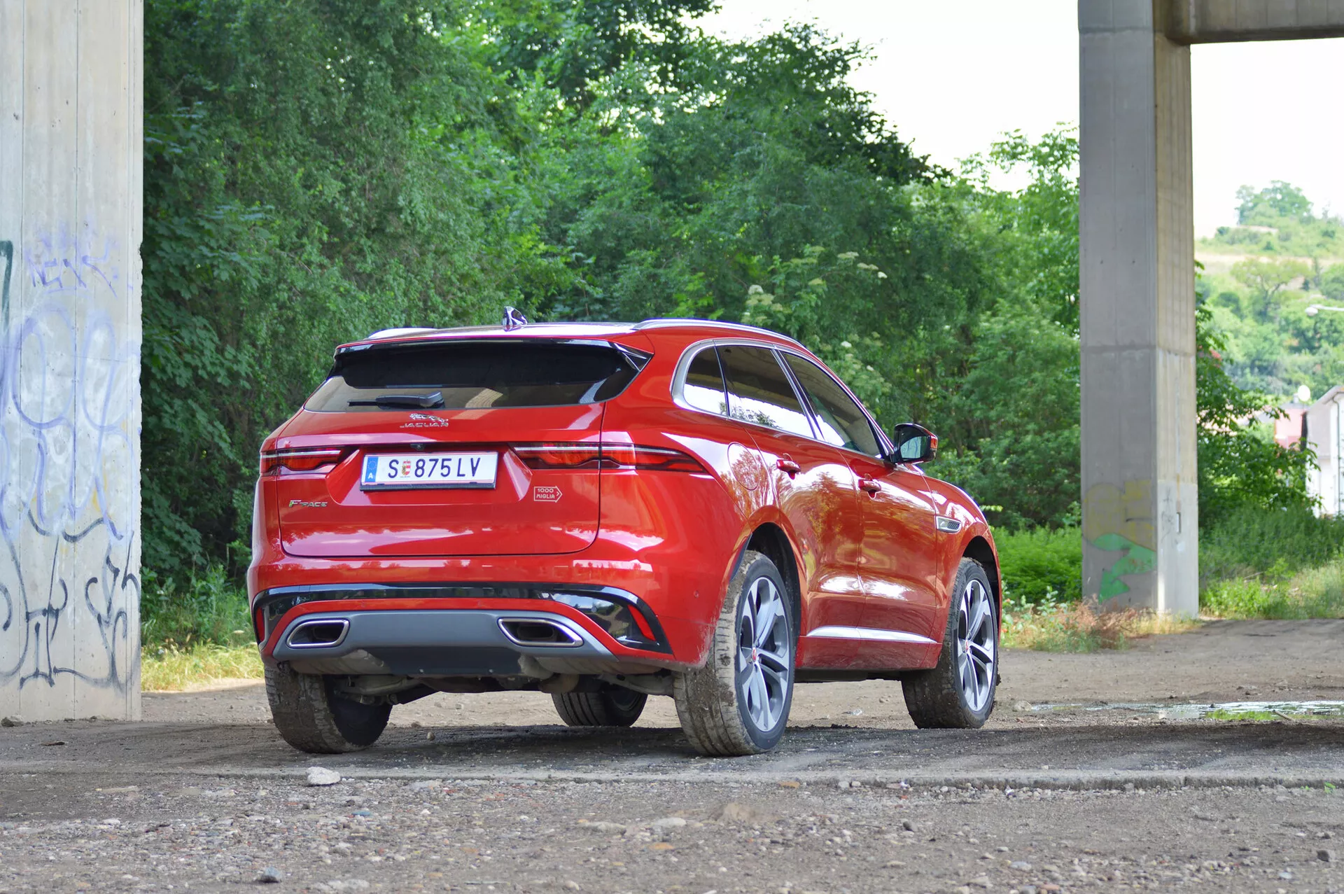 Trekvarts bakre vy av en röd 2021 Jaguar F-Pace SUV parkerad på en grov, oasfalterad yta med rester av gräs. Bilden visar bilens högra bakre sida och framhäver bakljusens design, bakre stötfångare och dubbel avgasrör, vilket antyder en sportig modell. En spoiler upptill på bakluckan och den sluttande taklinjen ger SUV:n ett coupe-liknande utseende. Miljön verkar vara urban, kanske under en bro, med betongstrukturer och graffiti i bakgrunden. Funktionerna inkluderar LED-bakljus, bakre parkeringssensorer och större hjul. Bilden är tagen på dagen i naturligt ljus.