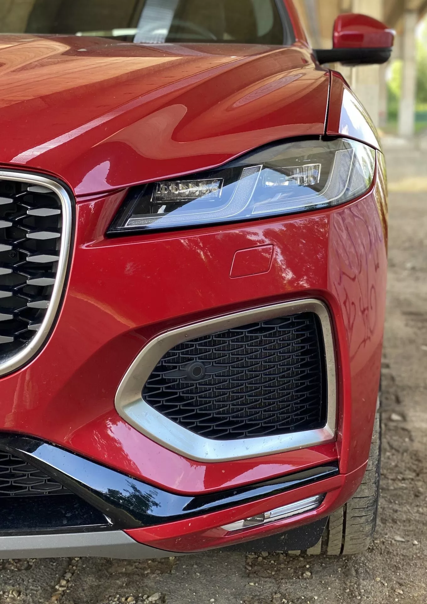 Närbild av den främre vänstra delen av en röd 2021 Jaguar F-Pace, med fokus på strålkastare, grill och främre stötfångare från en låg vinkel. Strålkastaren, med modern design och skarpa vinklar samt LED-belysningselement, och grillen med ett bikakemönster, är framträdande. Bilens blanka lack och aerodynamiska designlinjer betonas, med en svart kontrasterande sidobackspegel delvis synlig.