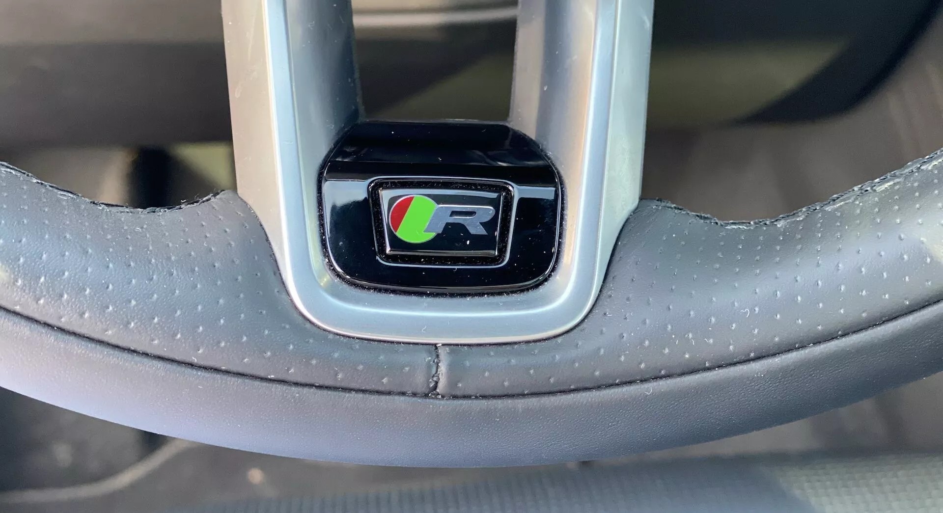 Närbild av rattens emblem på Jaguar F-Pace 2021 från förarens perspektiv. Emblemet, centrerat i bilden, är sköldformat med en distinkt 'R'-logotyp i svart med vit kontur mot en färgglad bakgrund i grönt, rött och blått. Emblemet är inneslutet i en blank svart inramning omgiven av en metallisk silverram. Den synliga delen av ratten är belagd med ett prickigt, greppförbättrande material, troligen perforerat läder eller liknande, vilket indikerar en lyxig och sportig design. Två horisontella ekrar sträcker sig från emblemområdet, men kontroller eller knappar är inte synliga.