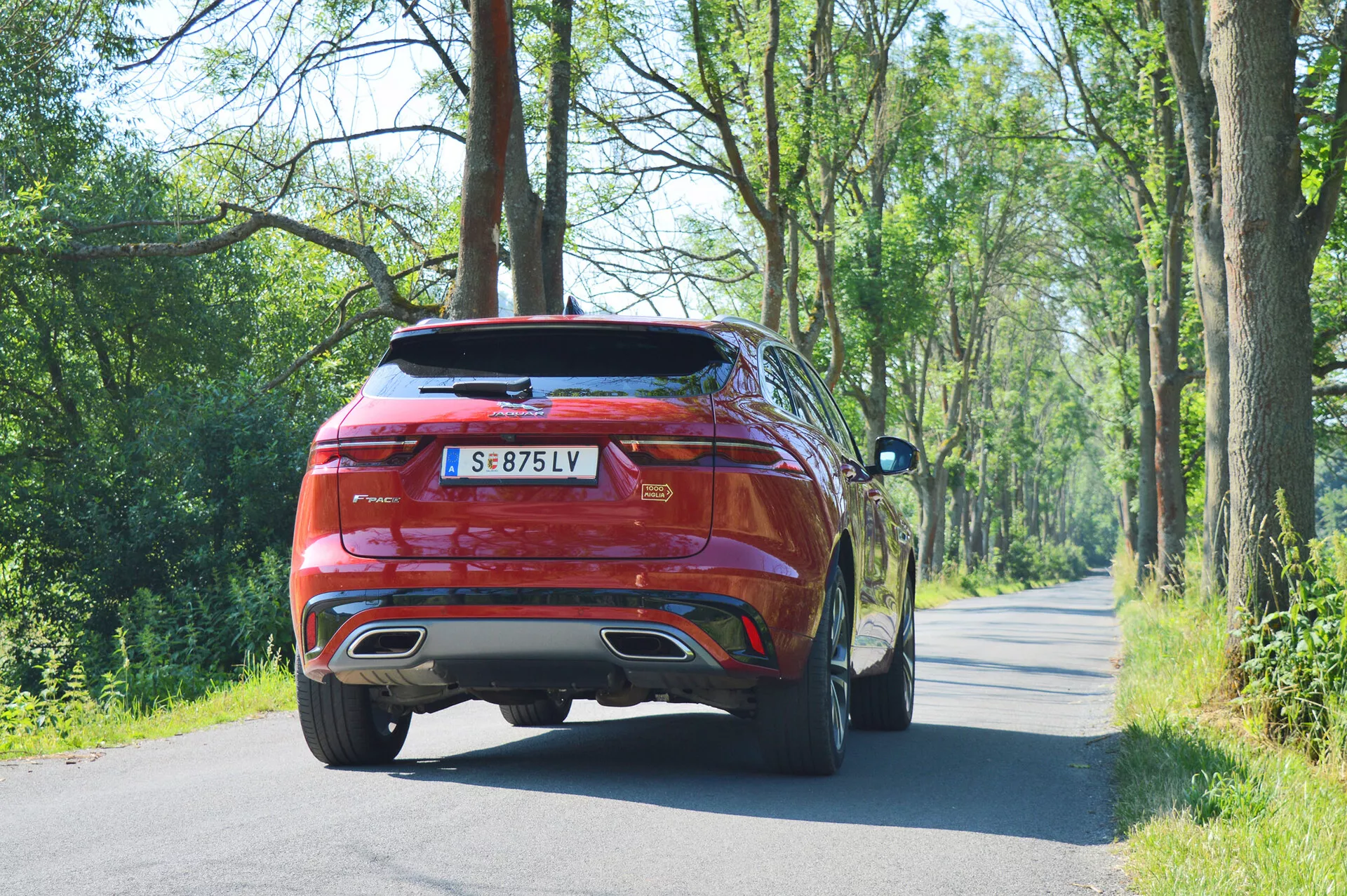 Bakvinkelvy av en mellanstor SUV Jaguar F-PACE 2021 på en väg omgiven av naturlig grönska. Bilen har en sportig, aerodynamisk design med sluttande taklinje, framträdande bakspoiler och horisontella LED-bakljus. Utrustad med stora lättmetallfälgar och dubbla avgassystem, har den en ljus metallisk röd lackering som indikerar en premium modell. Omgivningen antyder lämplighet för både stadskörning och landsbygdskörning.