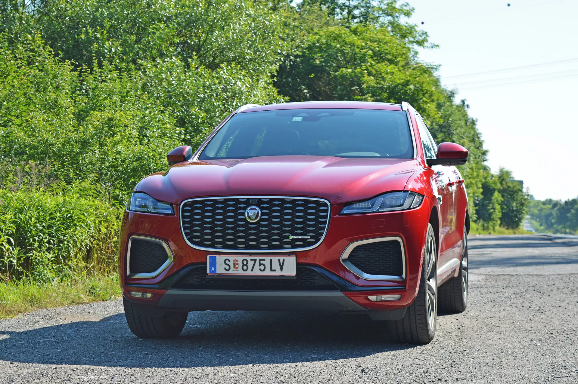 Frontal vinkelbild på en röd 2021 Jaguar F-Pace SUV tagen från en något lägre position, vilket betonar dess kraftfulla och imponerande design. Bilen upptar största delen av bilden och visar upp den stora främre grillen med en central logotyp, slanka LED-strålkastare, distinkta luftintag på den främre stötfångaren och en centralt monterad registreringsskylt. Synliga veck på den böjda huven förbättrar aerodynamiken. Bakgrunden innehåller lövverk och en klar himmel, vilket gör att bilens röda färg sticker ut tydligt.