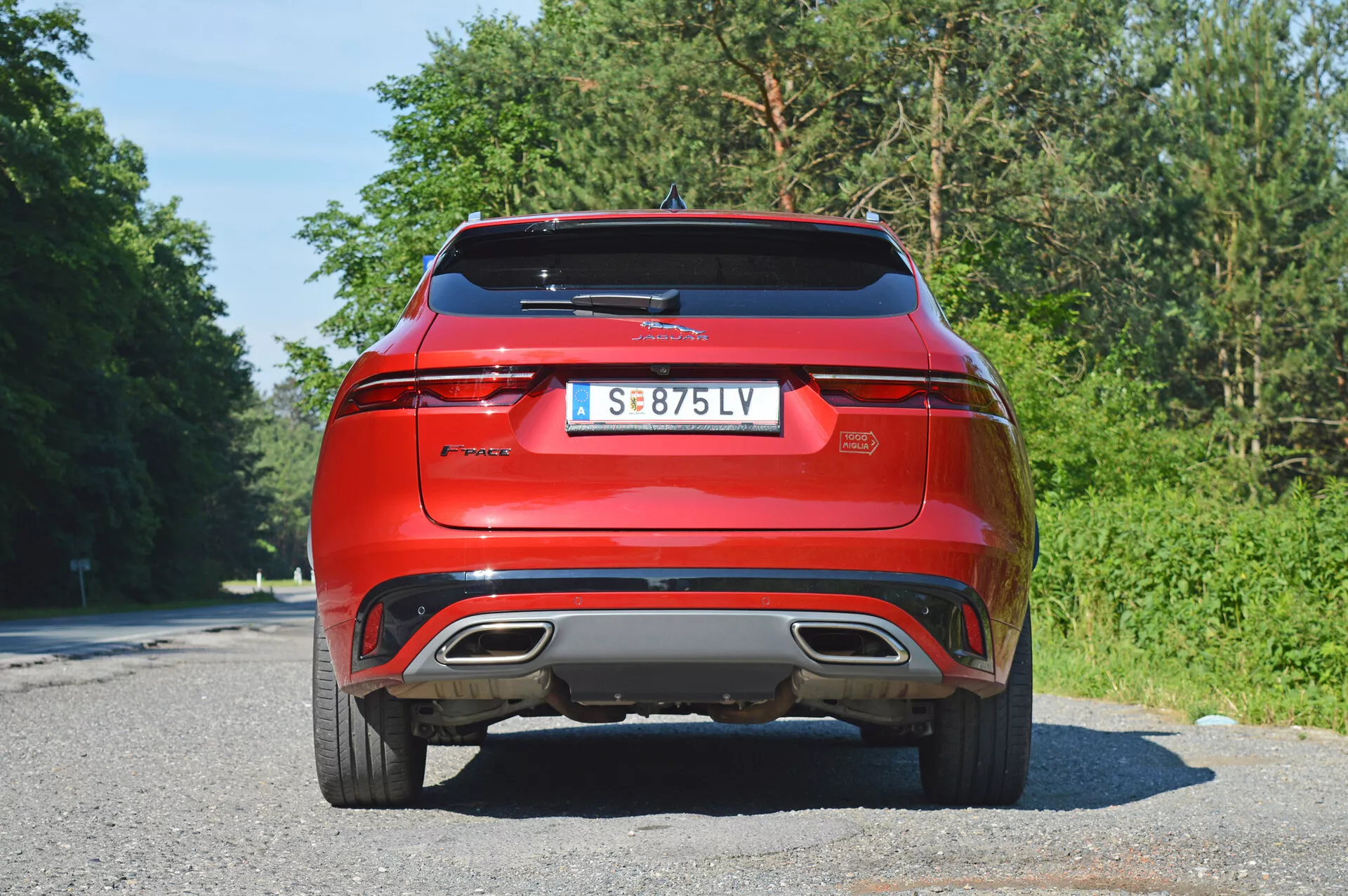 Bakre vy av röd Jaguar F-Pace 2021 parkerad på en asfaltsväg med träd i bakgrunden. Fotot är taget från en lätt lågvinkel som visar bilens moderna design med en böjd bakruta, integrerad spoiler, eleganta horisontella LED-bakljus, dubbla avgasrör, backkamera och hög bakdel, vilket antyder en sportig eller SUV-kaross.