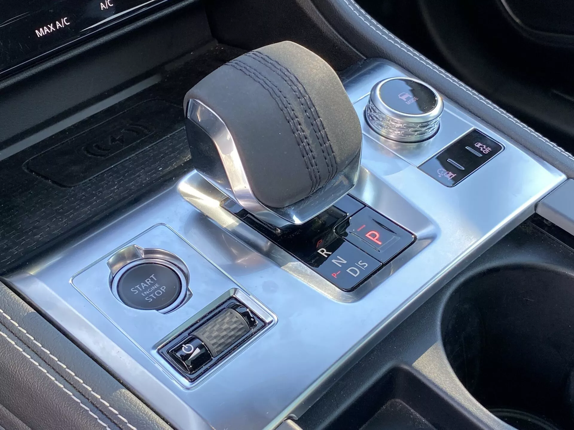 Detaljerad vy av området för mittkonsolen i 2021 års Jaguar F-Pace, med fokus på den elektroniska växelväljaren med läderliknande texturerad ratt, parkeringsindikator i rött, körlägesväljare och tangentförsedda motorstart/stopp-knappen. Funktioner inkluderar också parkeringsassistanssystemknapp och automatisk start-stop-system, omgiven av borstad aluminium-trim med synliga mugghållare och lyxig svart läderstygn.