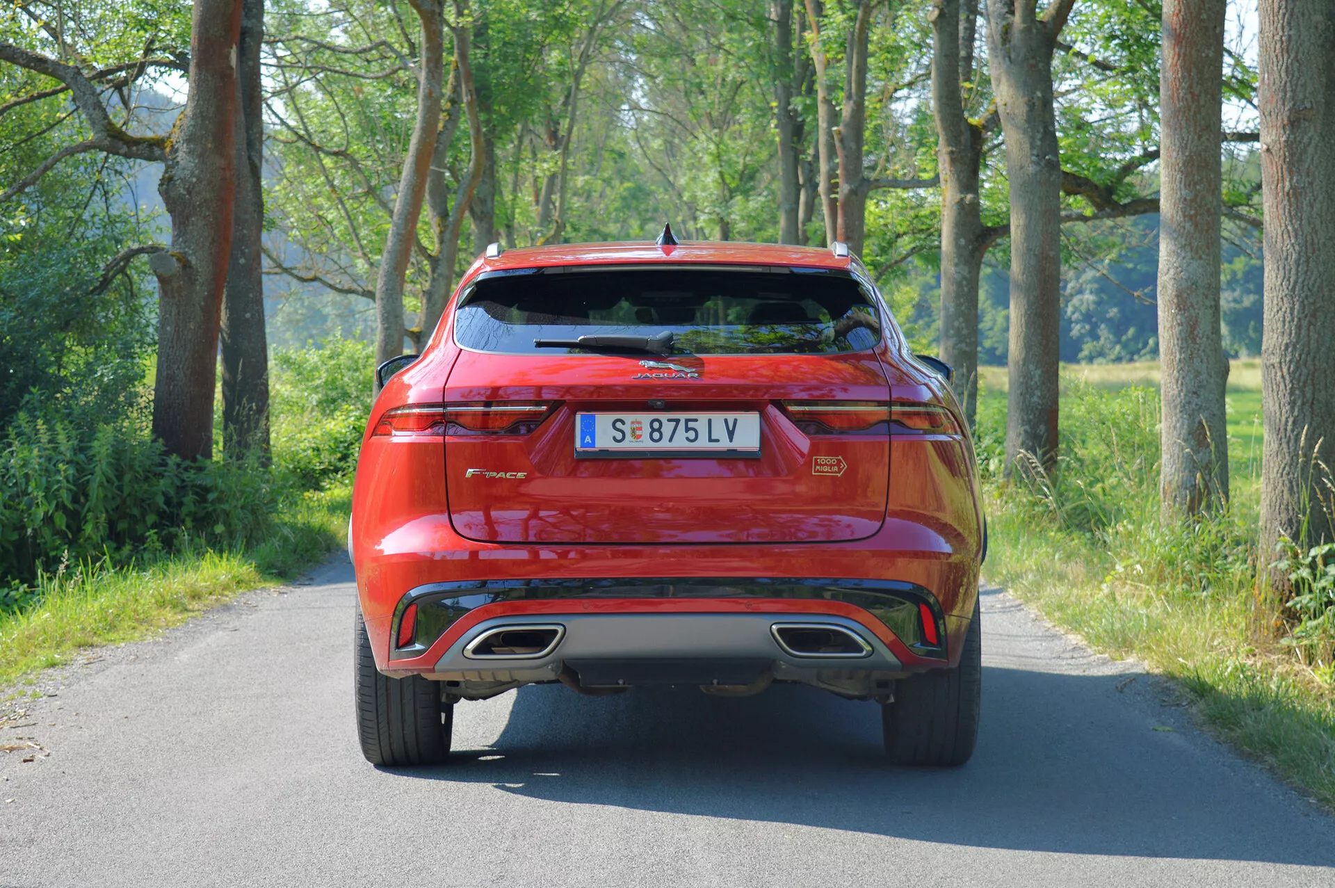 Bakifrån vy av en röd Jaguar F-Pace 2021 SUV på en smal, asfalterad landsväg, väl belyst under dagtid. Bilen har moderna LED-bakljus, modell- och märkesbadgering på bagageluckan, en centralt placerad registreringsskylt, dubbla avgasrör, en diffusordel på bakre stötfångaren, reflexer eller sensorer för säkerhets- och parkeringsassistans, en klar bakre vindruta med torkare och en blank lackfinish. Omgivningen antyder en lantlig eller halv-lantlig miljö.
