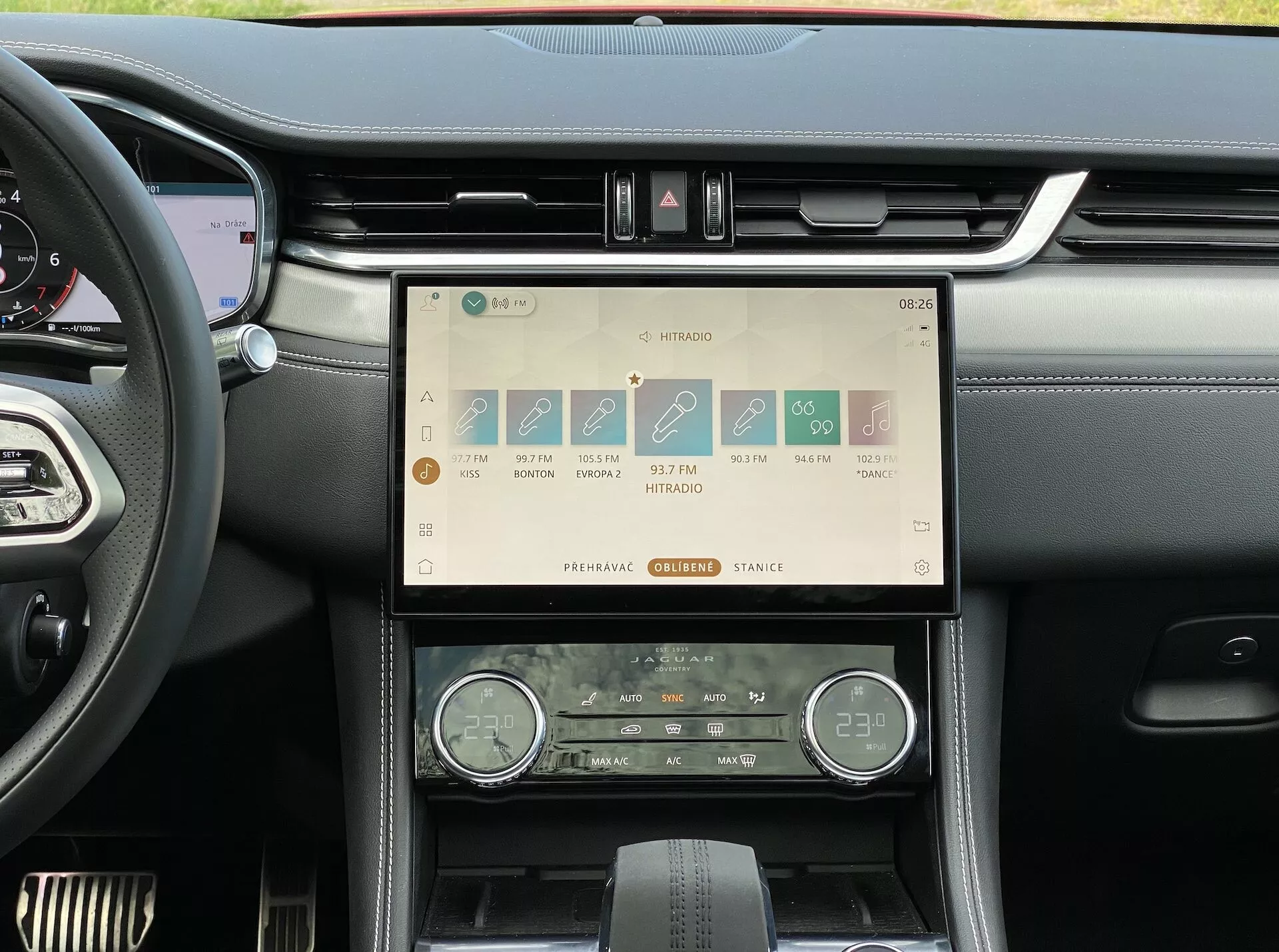 Interiörvy av instrumentbrädan i Jaguar F-Pace 2021 som visar mittkonsolen med ett stort, horisontellt orienterat pekskärmsinfotainmentsystem som visar färgglada ikoner för navigation, media, telefon och bilinställningar, tillsammans med en sekundär klimatanläggningsdisplay med en analog klocka, tvåzons temperaturer och fysiska kontrollreglage, vilket framhäver lyxiga material och elegant design.