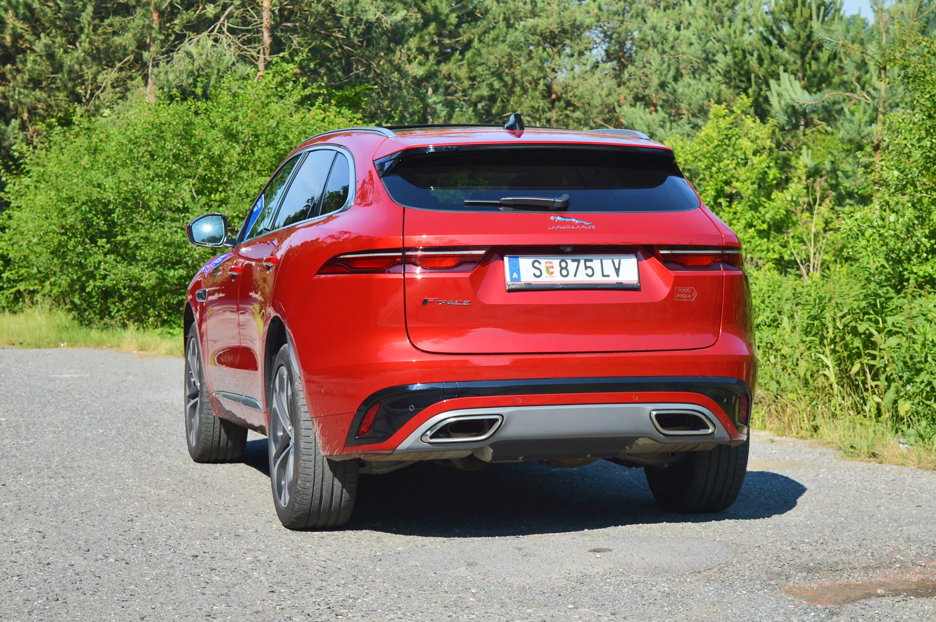 Bild av en röd Jaguar F-Pace 2021 fångad från en tredjedels vy bakifrån. Bilden är tagen från en lätt låg vinkel som framhäver fordonets eleganta horisontella LED-bakljus, den framträdande tillverkarlogotypen och modellmärket, dubbla integrerade avgasuttag, diffusorliknande bakre stötfångare med parkeringssensorer, avfrosterlinjer på bakrutan, liten takspoiler och fler-ekrade lättmetallfälgar. Bilen är parkerad vid vägkanten med växtlighet i bakgrunden, vilket antyder en lantlig eller förortsmiljö.