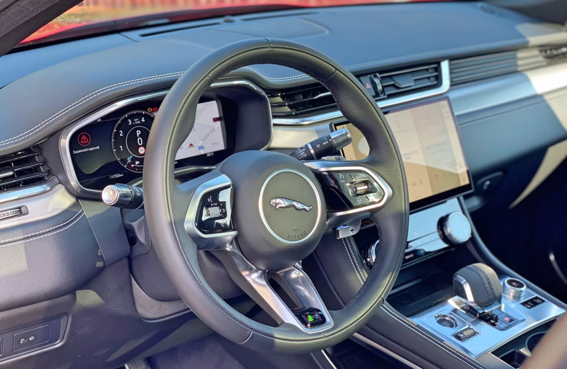 Interiörbild av en Jaguar F-Pace 2021 från förarsätets perspektiv, med fokus på ratten med tillverkarens emblem, en modern treekrad design med multifunktionskontroller, en digital instrumentpanel som visar förarfokuserad information, och en del av infotainmentsystemet som antyder avancerade teknologiska funktioner. Den centrala konsolen under pekskärmen inkluderar klimatkontrollknappar och en växelspak, vilket antyder en automatisk växellåda. Interiören har en blandning av material, möjligen med metalliska eller kromade accenter.