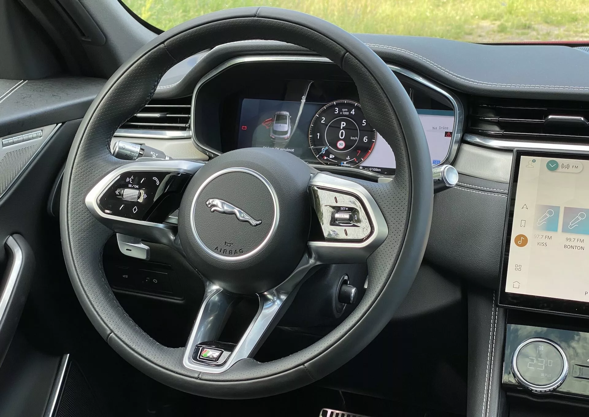 Interiörvy av Jaguar F-Pace 2021 från ett förstapersonsperspektiv, med fokus på ratten och instrumentbrädan. Den moderna treekrade ratten har kontroller och växelspakar som antyder möjligheten till manuell växling. Bakom ratten syns en digital instrumentpanel som visar hastighetsmätare och andra indikatorer, vilket potentiellt antyder en hybrid- eller eldriven drivlina. Till höger om ratten finns mittkonsolen med infotainmentsystemet som visar en karta, tillsammans med en klocka och klimatanläggningsknappar, vilket betonar lyxen av detta fordon.