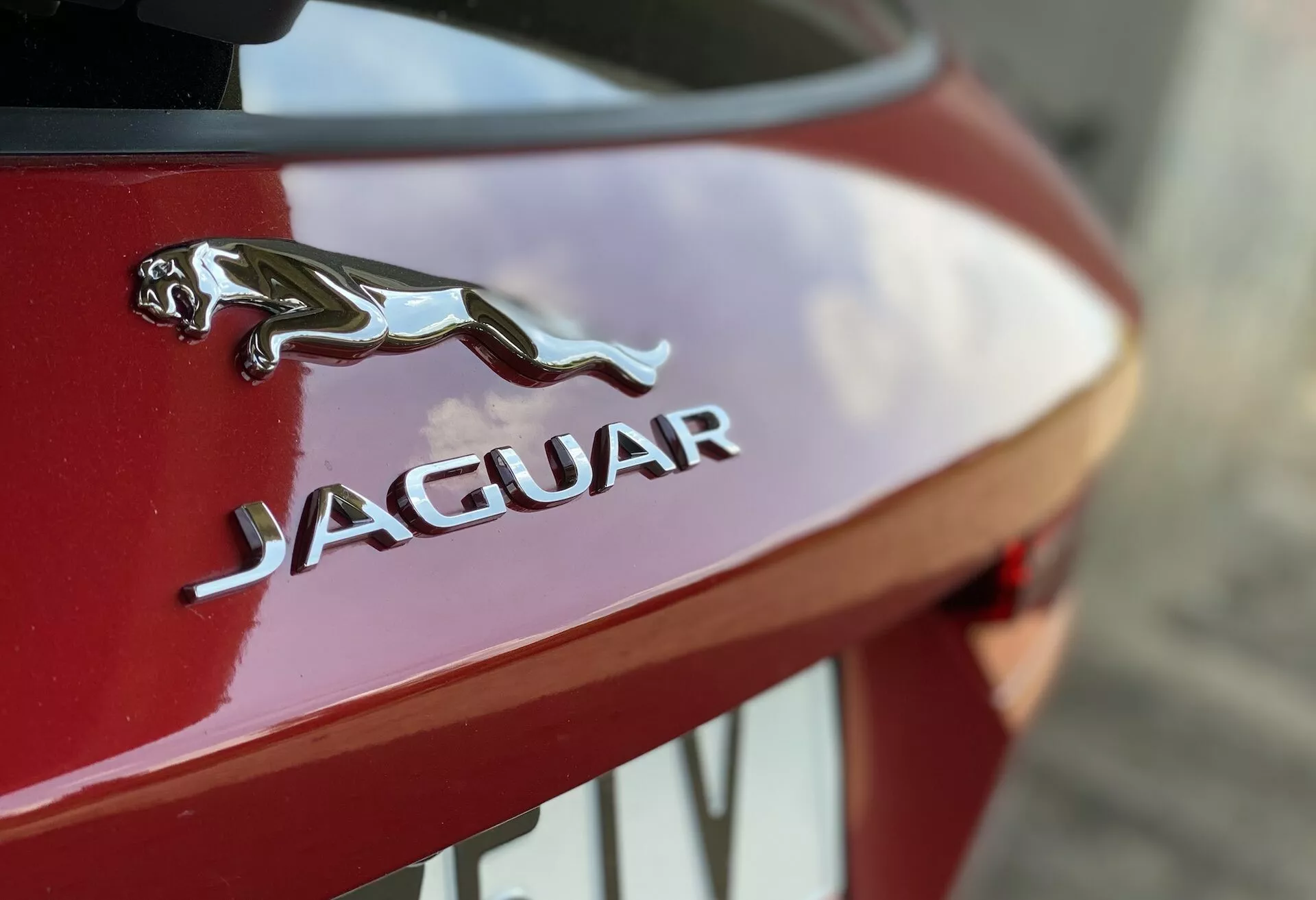 Närbild på bakre delen av en röd Jaguarbil, med kromad emblem av en hoppande Jaguar ovanför stiliserad 'JAGUAR'-text. Fotot fokuserar på denna varumärkesdetalj med en suddig bakgrund, vilket framhäver skarpheten i detaljerna, den blanka lacken och hantverket av lyxbilen.