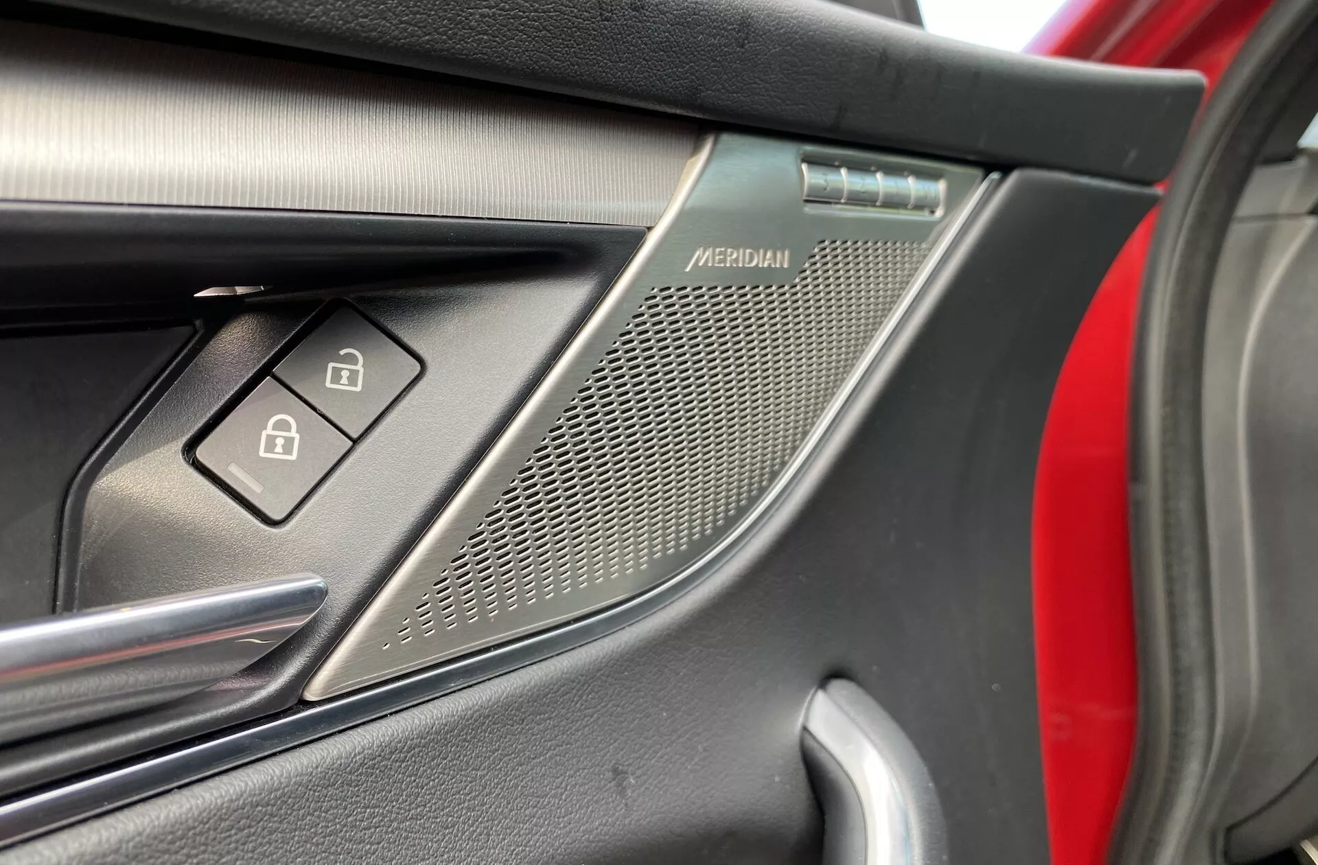 Närbild på förarsidans dörrinteriör i en Jaguar F-Pace 2021. Bilden fokuserar på ett Meridian-märkt högtalgaller i övre högra hörnet, spegeljusteringsknapparna nedanför och en del av dörrhandtaget. Dörrpanelen visar en blandning av material inklusive en metallisk kant och en texturerad insats. Den suddiga röda bakgrunden antyder bilens exteriörfärg.