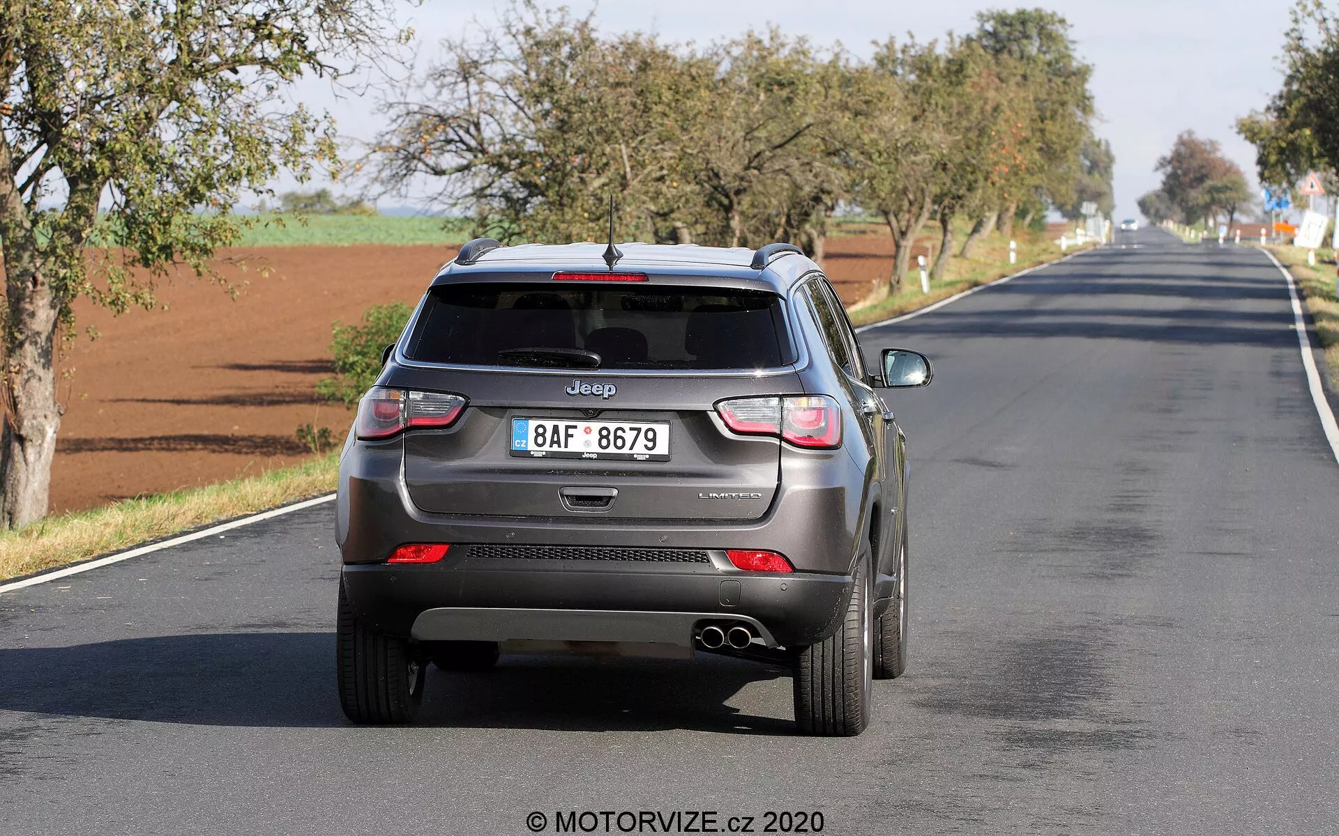 Bakifrånvy av en Jeep Compass 2017 som kör iväg på en tvåfilig väg, som visar moderna designelement som hajfensantenn, bakre vindrutetorkare, LED-bakljus, integrerade parkeringssensorer, dubbla avgasrör och en europeisk stil registreringsskylt med klart synlig Jeep-logotyp. Fordonet verkar vara i rörelse, vilket fångar verklig användning med potentiella förarstödteknologier.