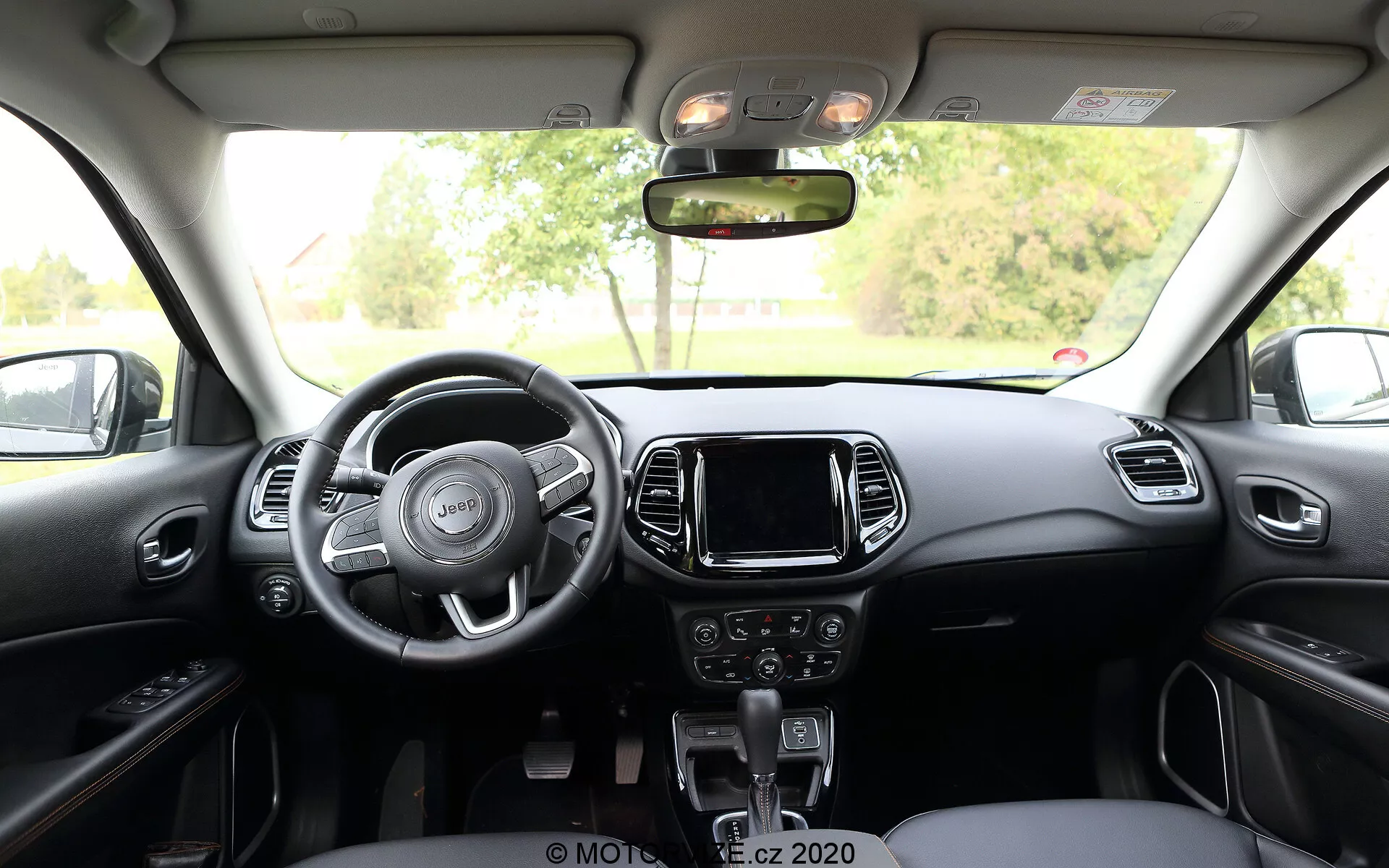 Interiörvy av 2017 års Jeep Compass från baksätet, som visar en treekrad ratt med inbyggda kontroller, mittkonsol med pekskärms-infotainmentsystem, manuella kontroller för ljud och klimat under den och en växelspak för automatisk växellåda. Instrumentpanelen och framsätena har en tvåfärgad design med metall- eller plastdekorationer, medan över huvudet finns innerbelysningsreglage och taklucka.
