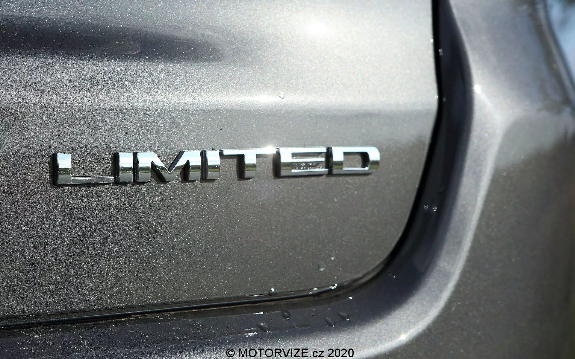 Närbildsdetalj av 2017 Jeep Compass Limited-märke med kromfinish på bakluckan; fotografiet fokuserar på märket från en vinkelperspektiv med en glänsande reflektion under direkt ljus, som suddar ut omgivande bildetaljer för att betona den högkvalitativa metallic-lacken.