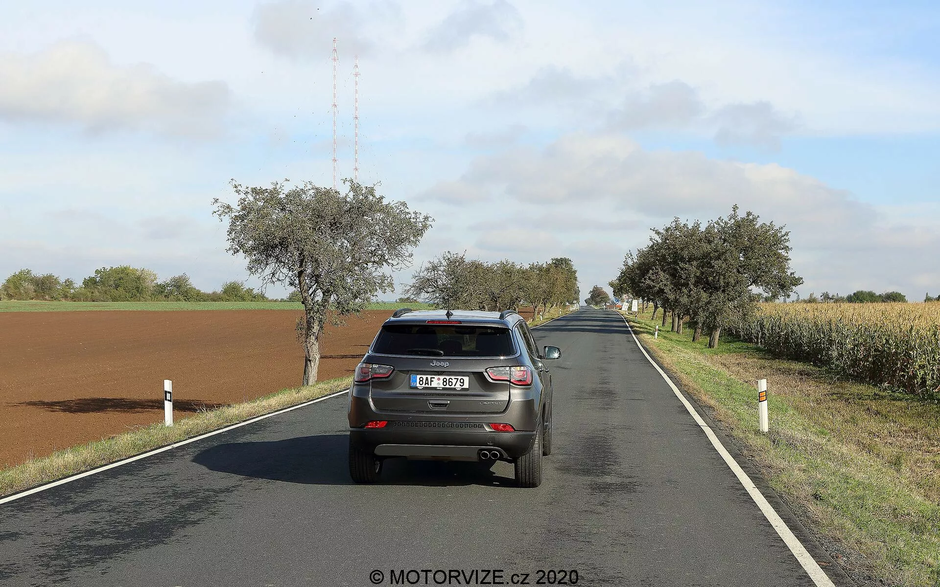Bilden visar bakåtvy av en 2017 Jeep Compass som kör på en tvåfilig landsväg på en solig dag med spridda moln. Fordonet, positionerat på höger sida av körfältet, visar sin baklucka, stötfångare, bakljus, bakruta, registreringsskylt, del av taket med hajfena-antenn och den övre delen av de bakre däcken. Bilen upptar en betydande del av ramen, vilket möjliggör tydlig detaljering av dess egenskaper. Vägen böjer sig mjukt åt höger i fjärran, omgiven av fält, träd och vägstolpar. Bilens bromsljus är inte tända, vilket tyder på normal körning.