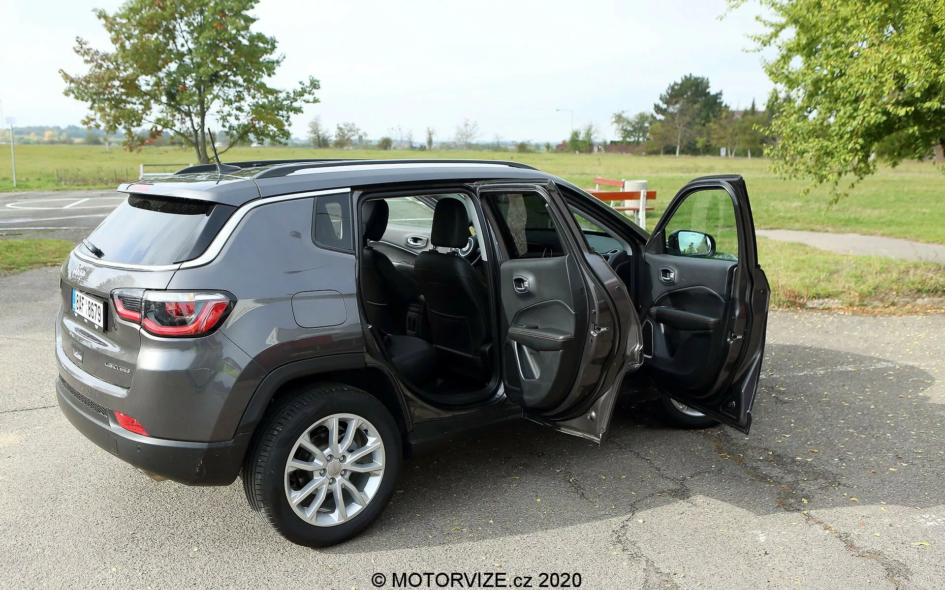 Bild av en 2017 Jeep Compass mellanstor SUV parkerad på en asfaltyta i en park-liknande miljö med gräs och träd i bakgrunden. Fotot är taget från en trekvarts bakvinkel som visar fordonets profil och bakände. Både passagerarsidans dörrar är öppna, vilket avslöjar den mörka tyg- eller läderinteriören inklusive fram- och baksäten, med möjliga funktioner som eldrivna fönster och speglar. SUV:en har en grå metallic-finish och är utrustad med fälgar i fem ekrar, vilket kan indikera en högre utrustningsnivå eller valfri hjuluppgradering. Vattenmärket 'MOTORVIZE.cz 2020' föreslår att bilden är från en bilrecension eller bilrelaterad webbplats.