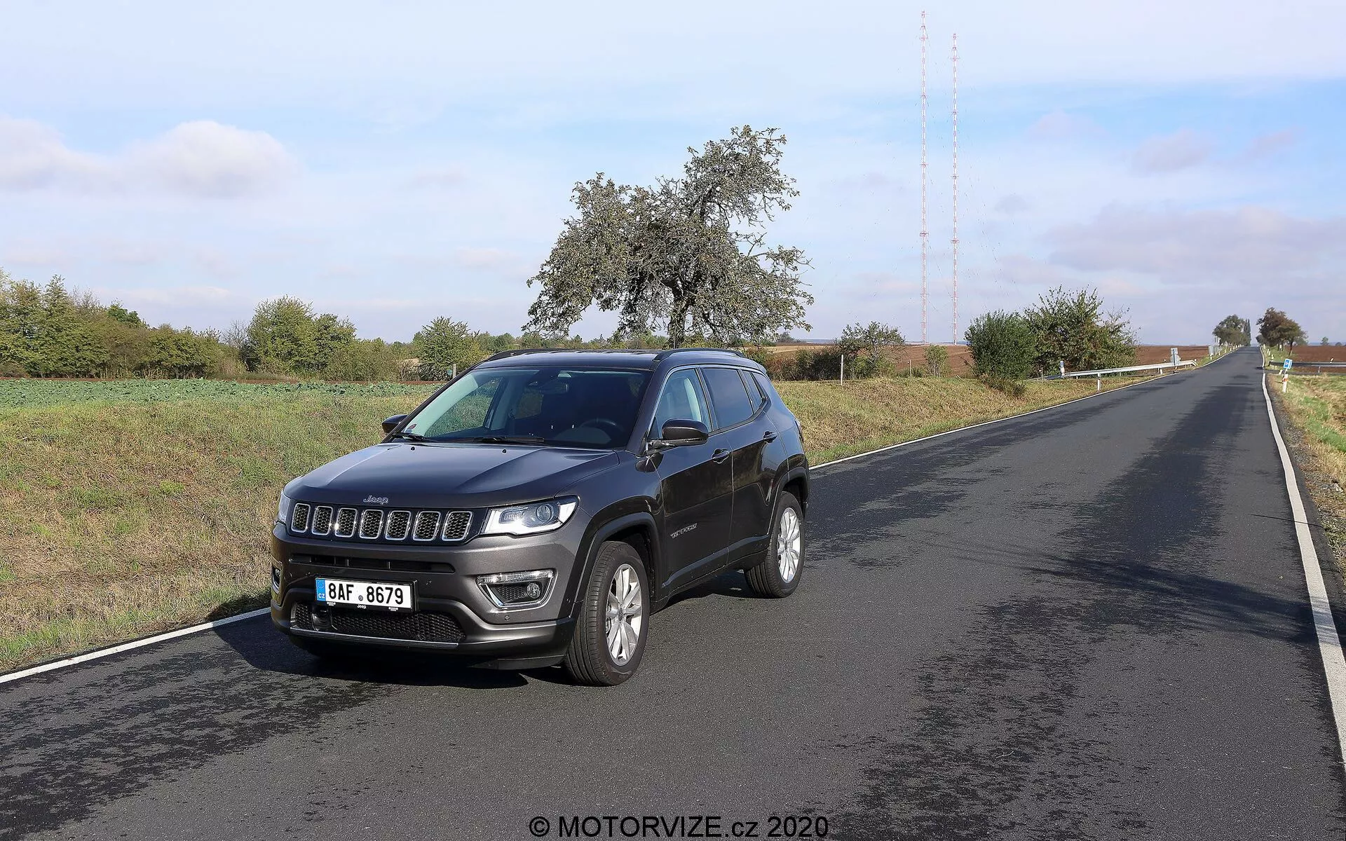 Bild på en Jeep Compass 2017 fotograferad från en trekvarts främre vinkel. Fordonet, placerat på höger sida av en tvåfilig väg, visar sin främre och sida profil. Nyckelfunktioner inkluderar en modern grill, framstående strålkastare med LED-dagljus, lättmetallfälgar, integrerade sidospegelblinkers, en takantenn och dimljus på den främre stötfångaren. Miljön verkar lantlig med klar himmel och växtlighet längs vägen.